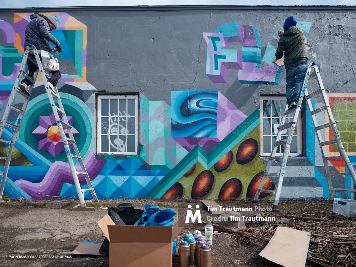 Two bundled artists perched on aluminum ladders collaborate on a vibrant geometric mural across a weathered gray concrete wall in Northeast Portland, Oregon. The emerging artwork features interlocking geometric patterns in turquoise, purple, and orange hues, incorporating circular mandala motifs and dimensional shapes that flow around existing barred windows. Scattered spray paint cans and cardboard boxes on the debris-strewn ground below capture the raw, industrial atmosphere of this urban art creation in progress.
