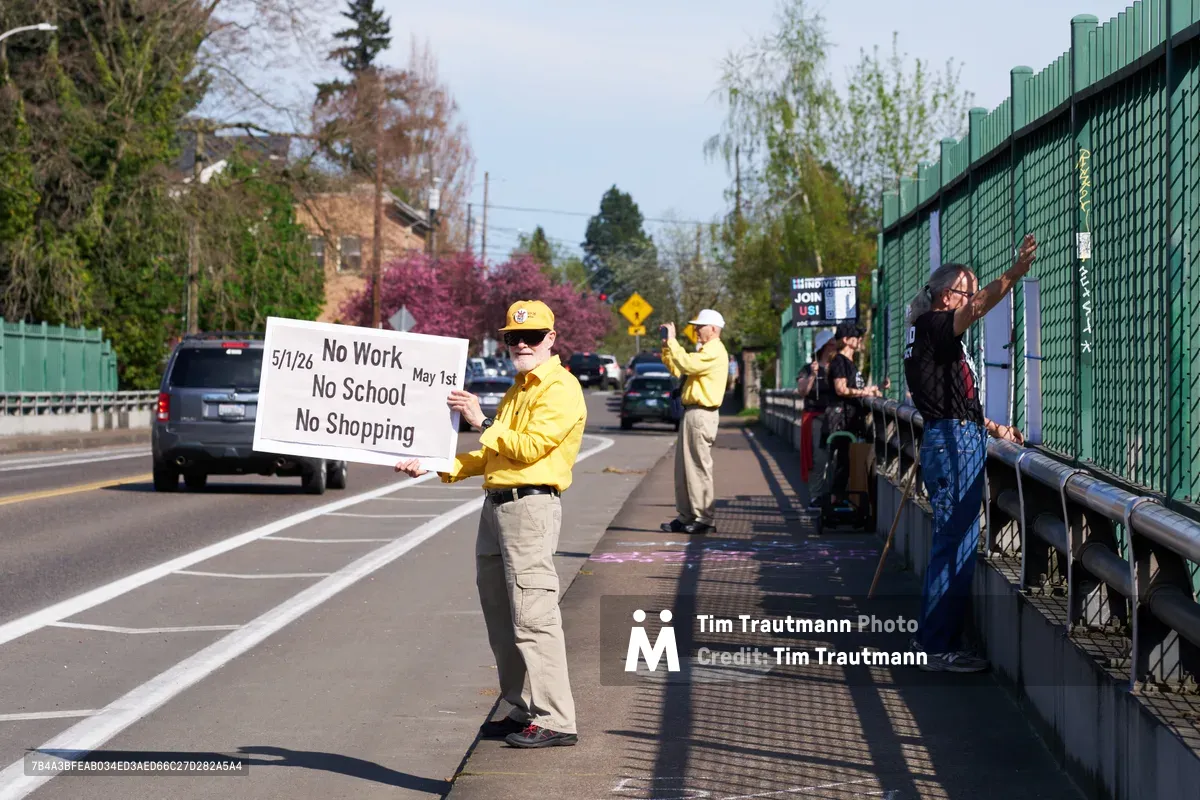 A determined activist in a bright yellow jacket and cap holds a handwritten protest sign reading "No Work No School No Shopping" while standing on North Skidmore Street's sidewalk above Interstate 5 in Portland. The late afternoon light casts long shadows across the concrete as fellow demonstrators gather along the green chain-link fence barrier, with spring foliage and residential buildings creating a suburban backdrop. Traffic flows steadily below as the Indivisible movement participants stage their May 1st General Strike awareness campaign.