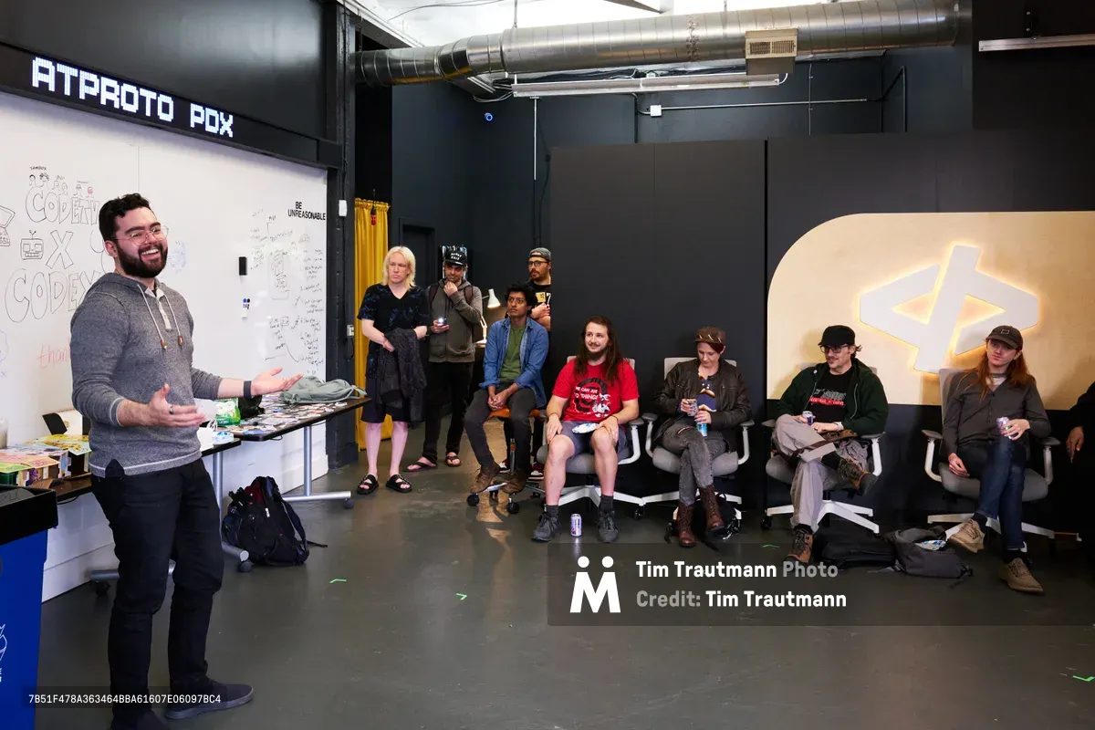 Graham, a bearded presenter in a gray sweater, gestures enthusiastically while addressing an intimate gathering of developers and tech enthusiasts seated in modern office chairs. The industrial Portland workspace features exposed ductwork, dark walls, and a prominent whiteboard covered in collaborative sketches and notes. Warm artificial lighting illuminates the engaged audience of eight attendees, creating an atmosphere of focused learning and community connection in this Ladd's Addition tech hub.
