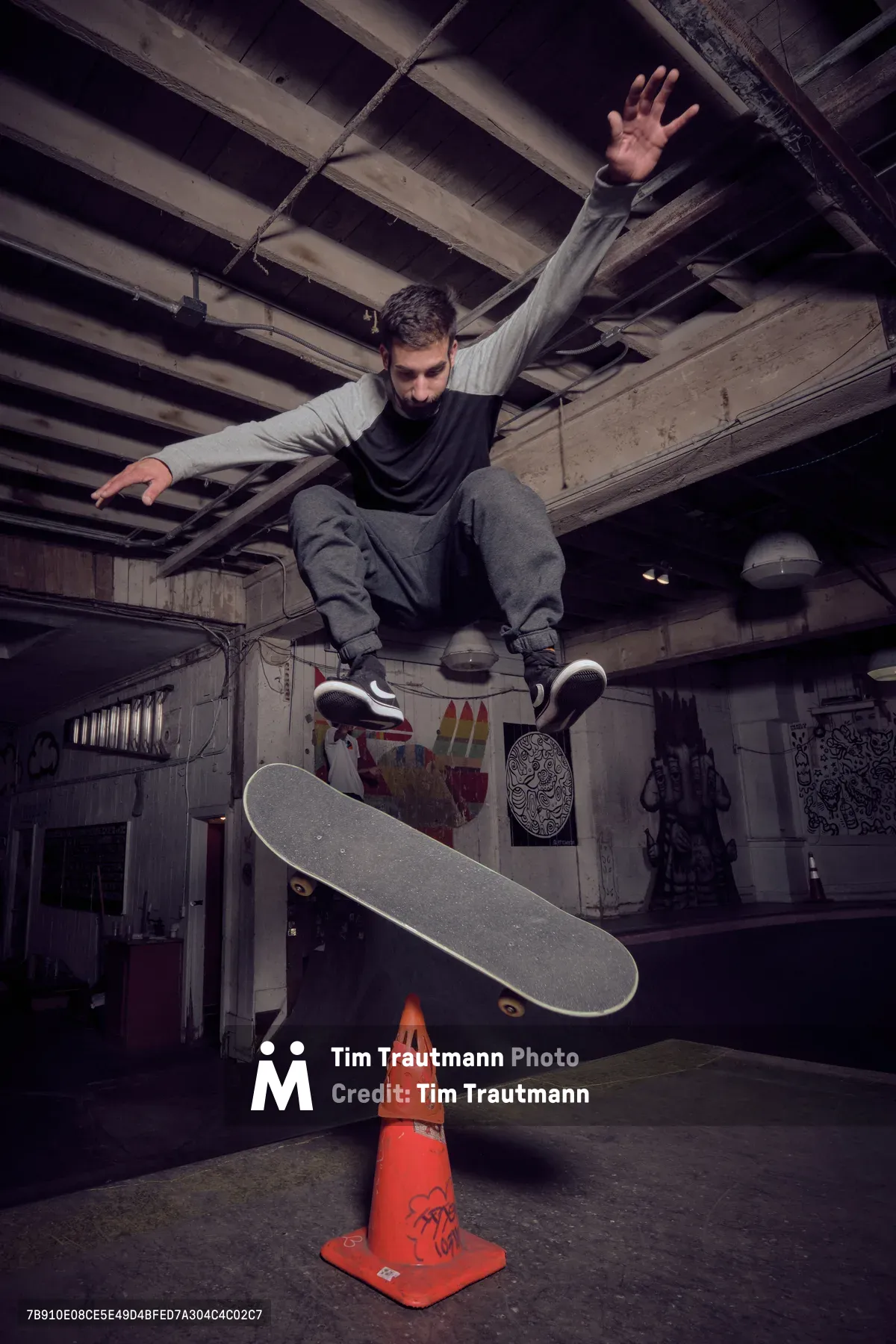 A bearded skateboarder launches into a gravity-defying ollie over a graffitied orange traffic cone atvCommonwealth Skateboarding in Portland's Buckman neighborhood. The moody lighting illuminates weathered wooden ceiling beams and concrete walls adorned with street art, while the athlete's arms spread wide in perfect aerial form. The underground venue's raw atmosphere creates a compelling contrast between urban grit and athletic grace.