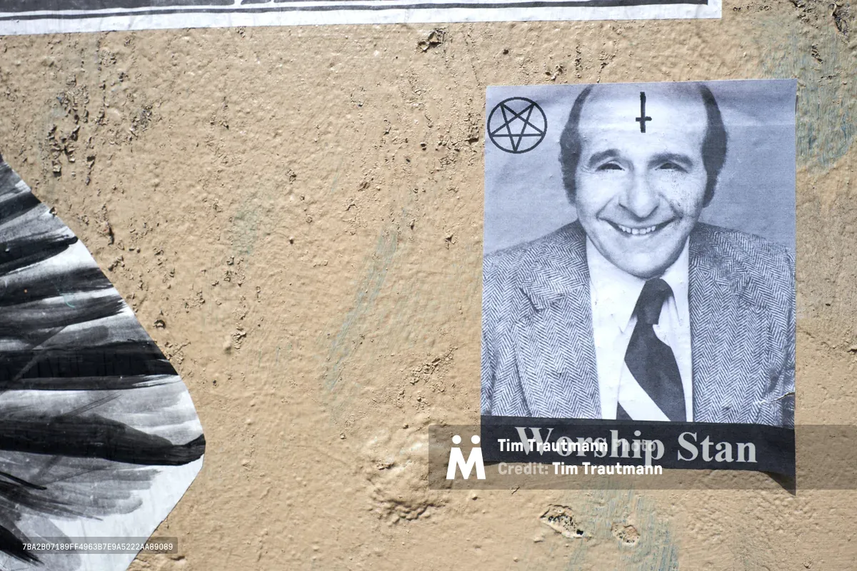 A provocative wheat paste poster adhered to a weathered concrete wall in Portland's Central Eastside features a black and white portrait of a smiling man in a suit and tie, adorned with occult symbols including a pentagram and inverted cross. The text "Worship Stan" appears beneath the portrait in bold white letters against a black banner. Adjacent torn paper fragments create textural contrast against the gritty urban surface, capturing the raw aesthetic of guerrilla street art in the Grand Avenue Historic District.