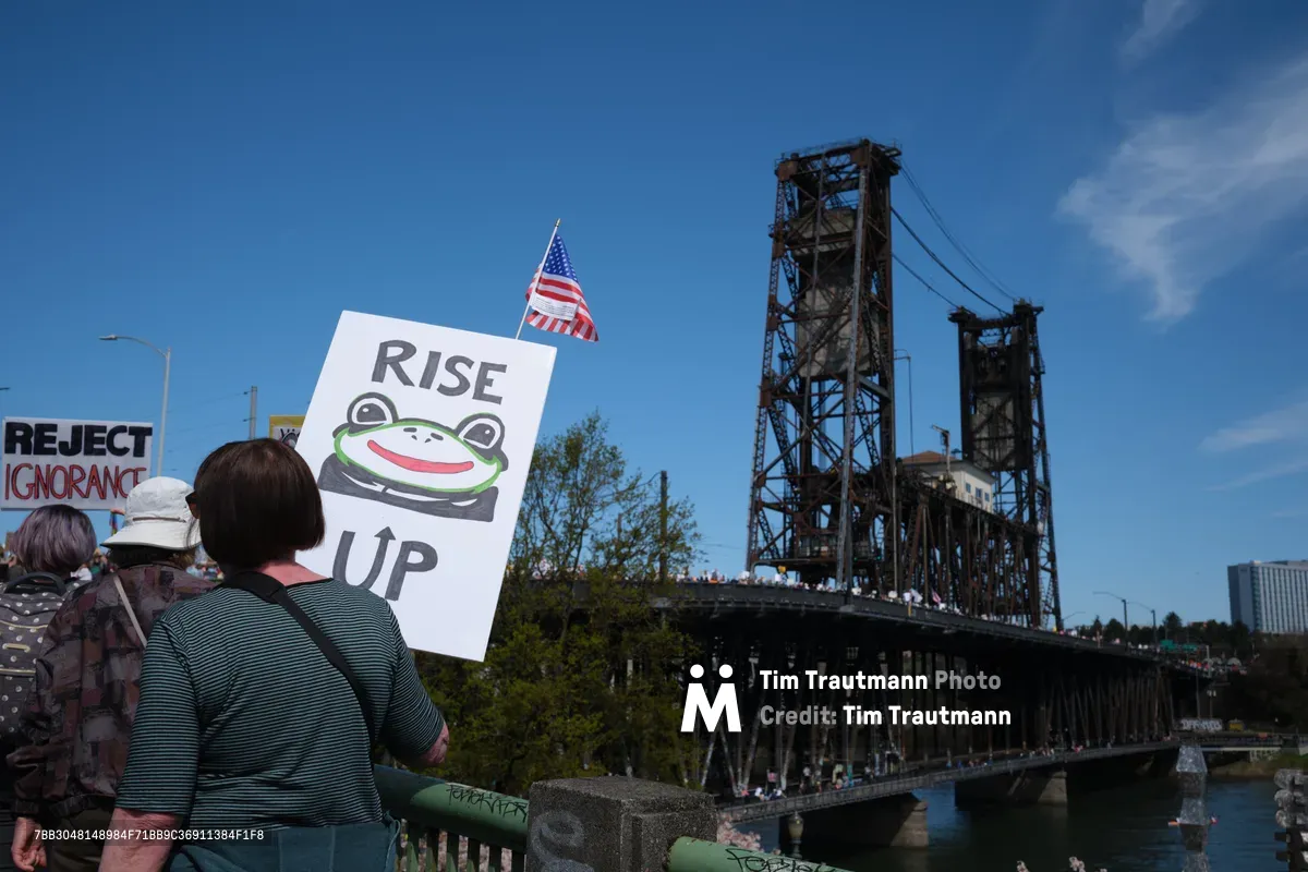 Demonstrators gather beneath Portland's iconic Steel Bridge during the March 2026 No Kings Protest, their signs and American flag silhouetted against a brilliant azure sky. A protester holds a distinctive 'RISE UP' placard featuring Pepe the Frog imagery, while the weathered steel lattework of the historic drawbridge looms overhead like an industrial cathedral. The scene captures a moment of civic engagement framed by the urban landscape of the Lloyd District, where political expression meets architectural heritage under the Pacific Northwest's crystalline light.