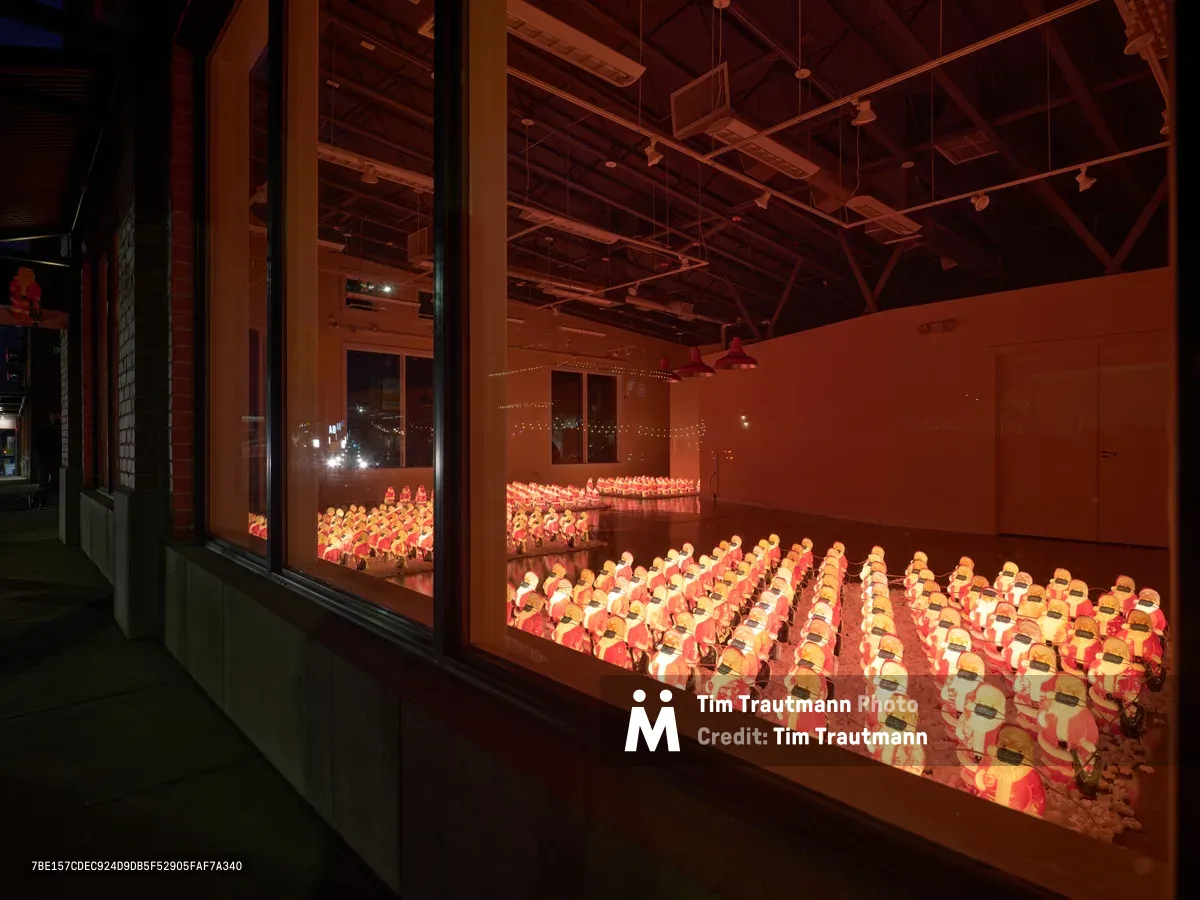 Hundreds of identical Santa Claus figures wearing protective face masks fill tiered seating in a Portland venue, their warm orange glow contrasting against exposed industrial ceiling beams. The installation by Chris Willis transforms the familiar holiday figure into a poignant commentary on collective isolation during the COVID-19 pandemic. Viewed from an elevated corridor through floor-to-ceiling windows, the repetitive figures create a mesmerizing pattern that speaks to both unity and distance in unprecedented times.
