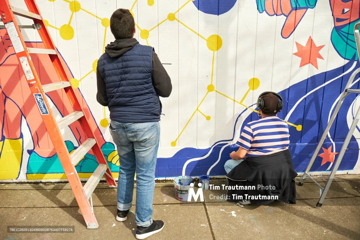 Two muralists collaborate on 'Let's Talk,' a vibrant community artwork at Open Signal in Portland's historic Eliot neighborhood. One artist, wearing a navy vest and jeans, observes the geometric yellow network pattern while their colleague in a blue striped shirt sits cross-legged on the concrete, headphones on, painting flowing blue and coral forms. An orange stepladder stands ready against the white brick wall, paint cans scattered nearby as the mural takes shape under natural daylight.