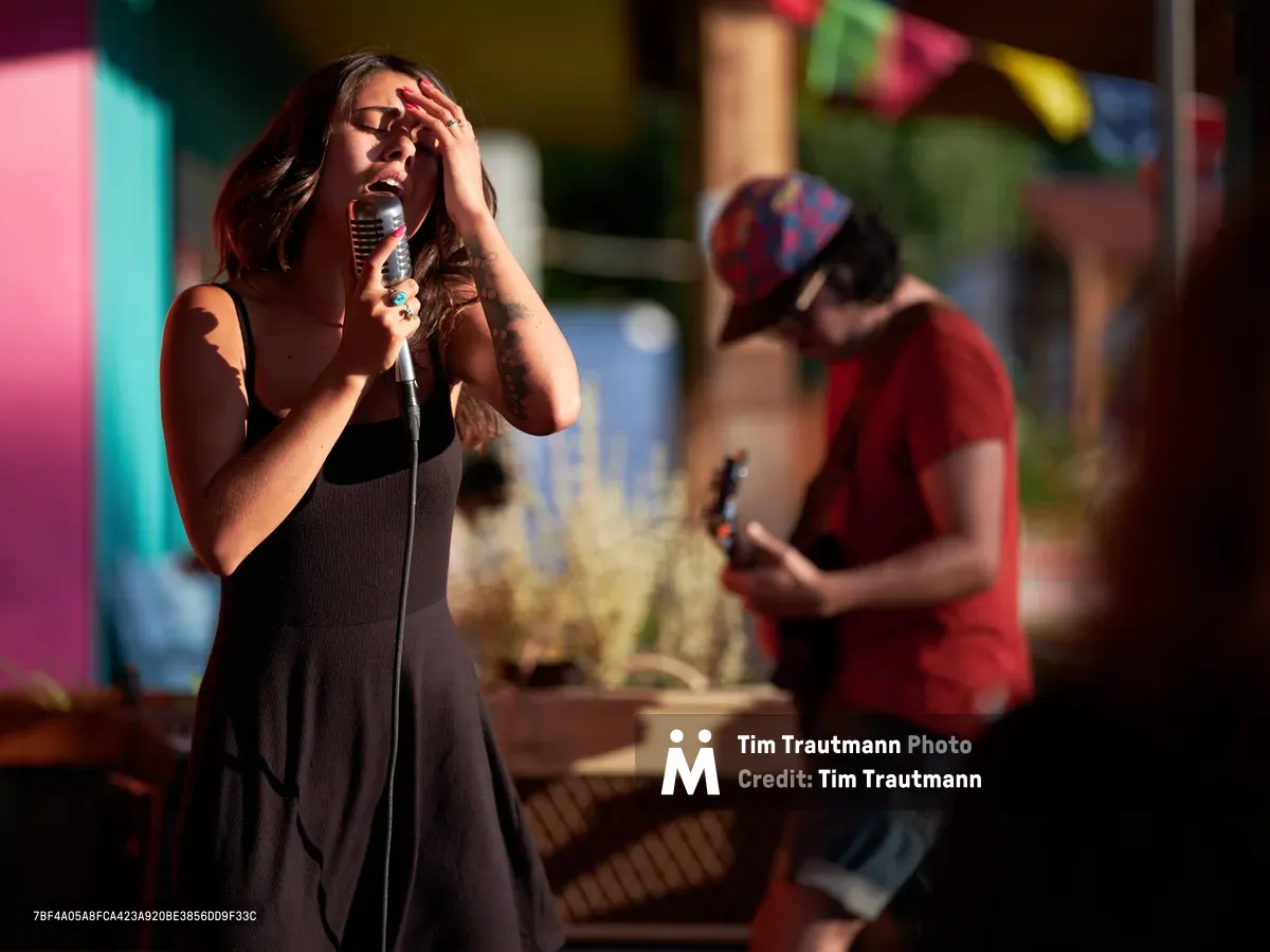 Aubrey Gordon loses herself in song during an intimate performance at Portland Mercado, her hand pressed dramatically to her forehead as golden sunlight bathes the scene. The vintage microphone captures her passionate delivery while a guitarist in a red shirt provides accompaniment in the softly blurred background. The warm evening light creates a nostalgic atmosphere against the colorful market setting of Southeast Portland's beloved food cart pod.