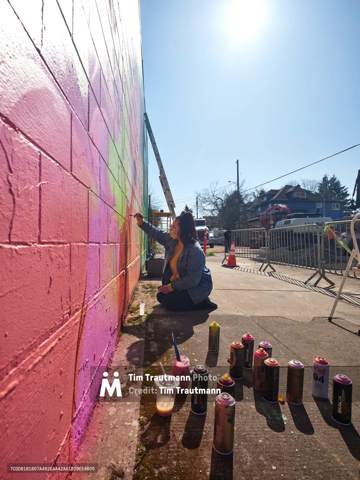 Multidisciplinary artist Villatose kneels on Northeast Alberta Street's sidewalk, applying bold magenta and lime green spray paint to create 'Jardín Cubana,' a mural featuring three female figures on a concrete wall. Dozens of aerosol cans scatter across the pavement in a kaleidoscope of colors, while construction barriers frame this creative transformation in Portland's celebrated Alberta Arts District. The afternoon light catches the fresh pigment, creating an intimate documentation of street art in progress.