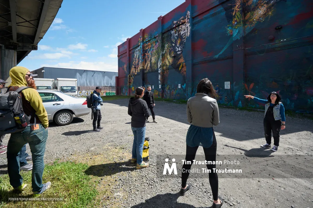 A diverse group of participants gathers on weathered asphalt beneath spring's clear blue sky, following tour guide Tiffany Conklin as she gestures toward vibrant murals adorning the industrial walls of Portland's Central Eastside. The scene captures the intersection of urban art and community engagement, where concrete infrastructure meets colorful creative expression. Shadows fall sharply across the gravel lot, while participants in casual attire crane their necks to absorb the cultural narratives painted across the warehouse facades.
