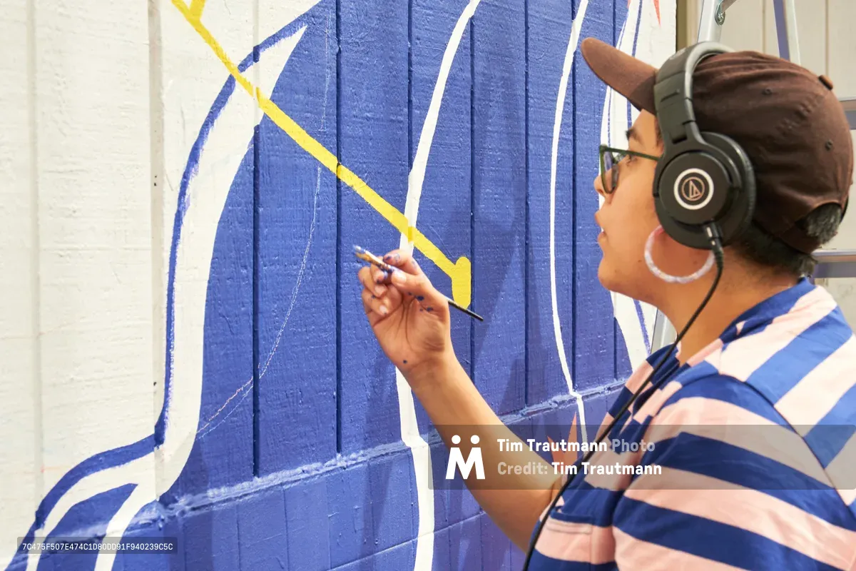Maria Rodriguez, known as Sparkykneecap, works intently on a vibrant blue and yellow mural titled "Let's Talk" at Open Signal in Portland's Eliot neighborhood. Wearing headphones and a blue-striped shirt, the Mexican-American artist carefully applies paint to the wooden fence surface in bright afternoon light. The geometric composition captures the meditative focus of creation, with bold cerulean blues and sunny yellows forming abstract shapes that speak to themes of identity and cultural connection.
