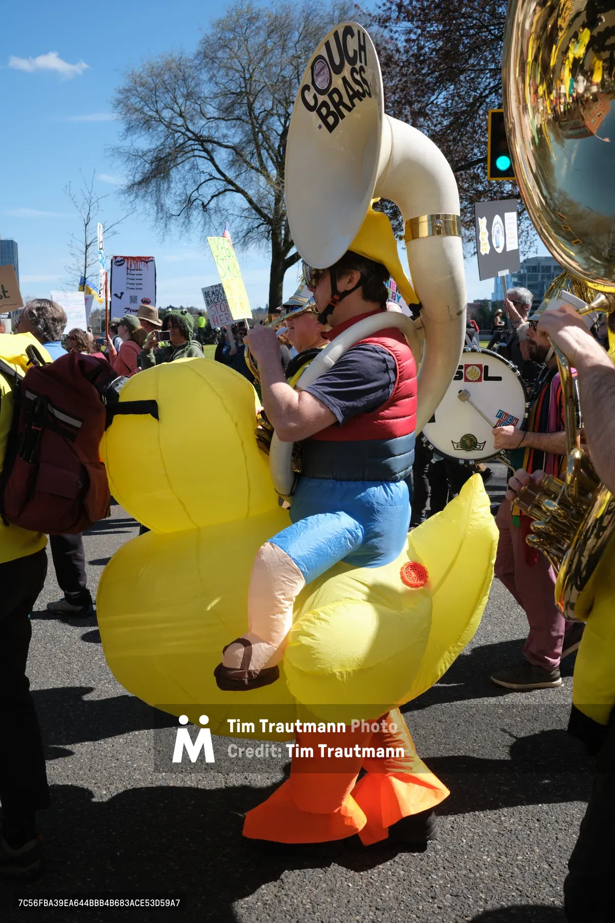 A tuba player wearing an inflatable yellow rubber duck costume performs with the Unpresidented Brass Band during a protest march in Portland's Old Town district. The musician, strapped into the whimsical costume with orange webbed feet, plays beneath bare spring trees while surrounded by fellow protesters carrying signs. The scene captures the intersection of political activism and performance art, with bright afternoon sunlight illuminating the colorful spectacle against the urban backdrop of Southwest Ankeny Street.