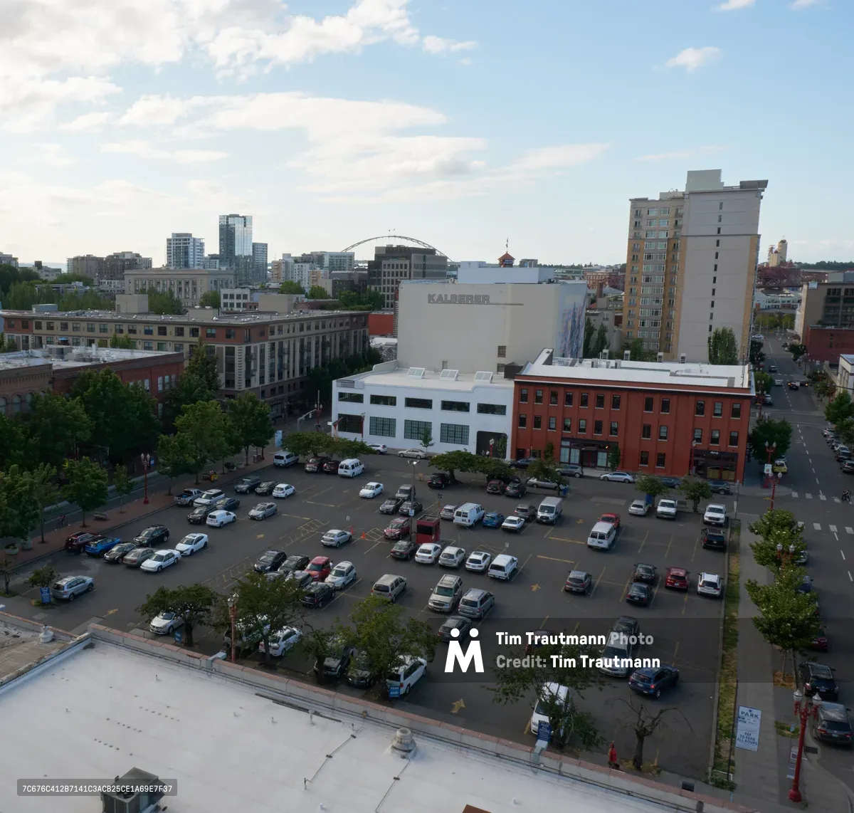 An elevated view captures the diverse architectural tapestry of Portland's Old Town district, where a surface parking lot serves as the immediate foreground beneath gathering afternoon clouds. The scene reveals the characteristic blend of historic red brick buildings and contemporary structures that define this Pacific Northwest neighborhood, with the Kalberer building prominently anchoring the middle ground. Soft natural light filters through a partly cloudy sky, casting gentle shadows across the urban landscape and creating depth between the foreground parking area and the distant high-rise towers.