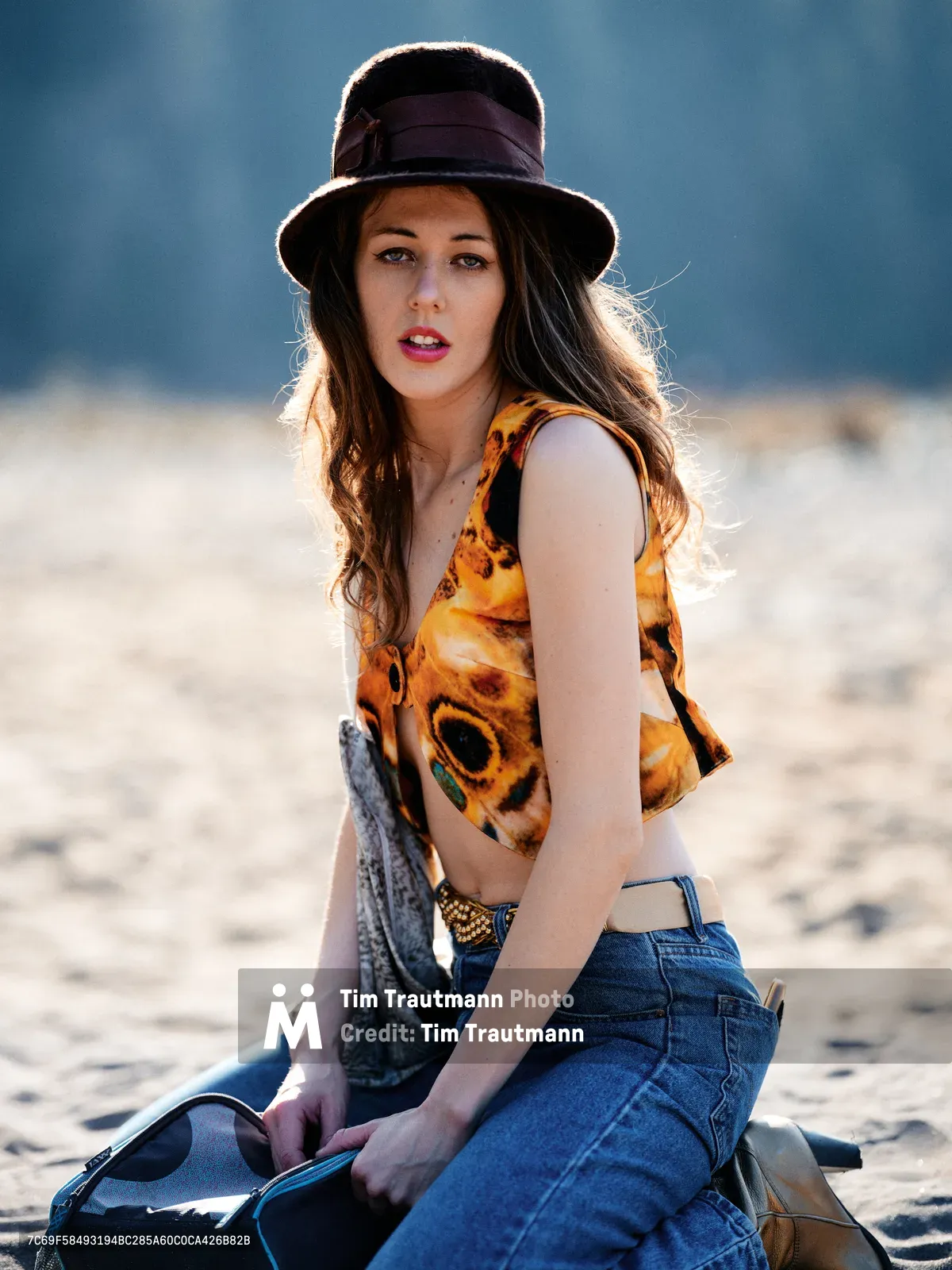 A young woman sits contemplatively on sandy riverbed terrain along Oregon's Floodplain Trail, wearing a distinctive purple fedora hat and vibrant orange crop top with abstract patterns. The golden hour backlighting creates a warm rim light around her brunette hair, while she holds what appears to be a denim jacket. Her direct gaze and relaxed posture convey confidence against the blurred natural landscape of Multnomah County's outdoor recreation area.