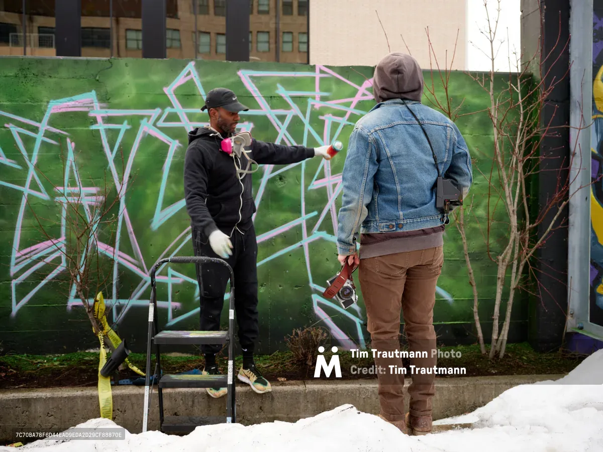 In the creative enclave of Southeast Portland's Hosford-Abernethy neighborhood, a street artist in black hoodie and cap applies spray paint to a vibrant geometric mural while engaging in conversation with a photographer in denim jacket and brown pants. The emerald green wall serves as canvas for angular white and pink lettering, with urban apartment buildings rising in the soft overcast light behind bare winter branches.
