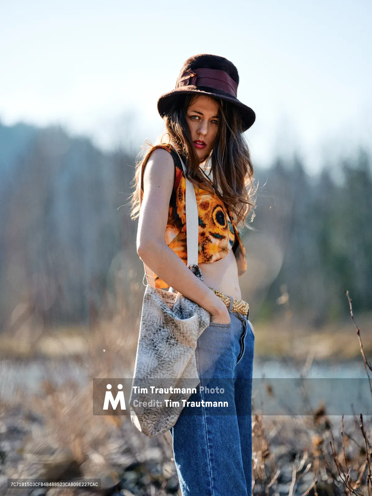A young woman with long flowing hair stands in contemplative pose along the Floodplain Trail in Multnomah County, Oregon. She wears a distinctive black wide-brimmed hat and layered bohemian attire including a graphic tank top and denim, while carrying a woven bag. The golden hour light creates a warm rim-lit effect against the soft-focus river bed landscape of dried grasses and distant water, with forested hills rising in the background.
