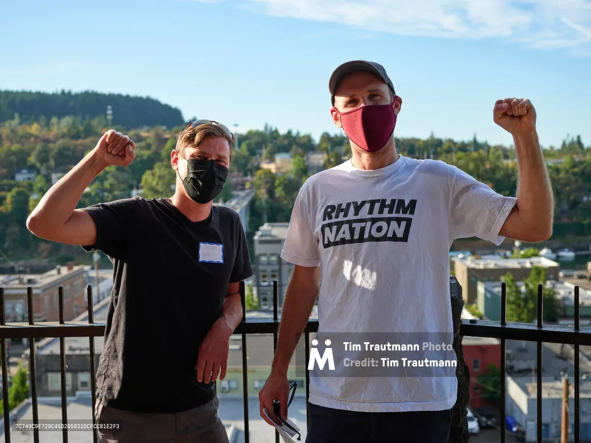 Michael Gaskill and Peter Marks raise their fists in solidarity against a sweeping vista of Oregon City's hillside neighborhoods, their masked faces reflecting the pandemic-era context of their climate activism. The afternoon light illuminates their determined expressions as they pose on the McLoughlin Promenade balcony, with the forested hills and residential sprawl of Clackamas County creating a layered backdrop. Gaskill wears a black shirt while Marks displays his commitment with a white "Rhythm Nation" tee, both men embodying the grassroots energy of environmental protest.