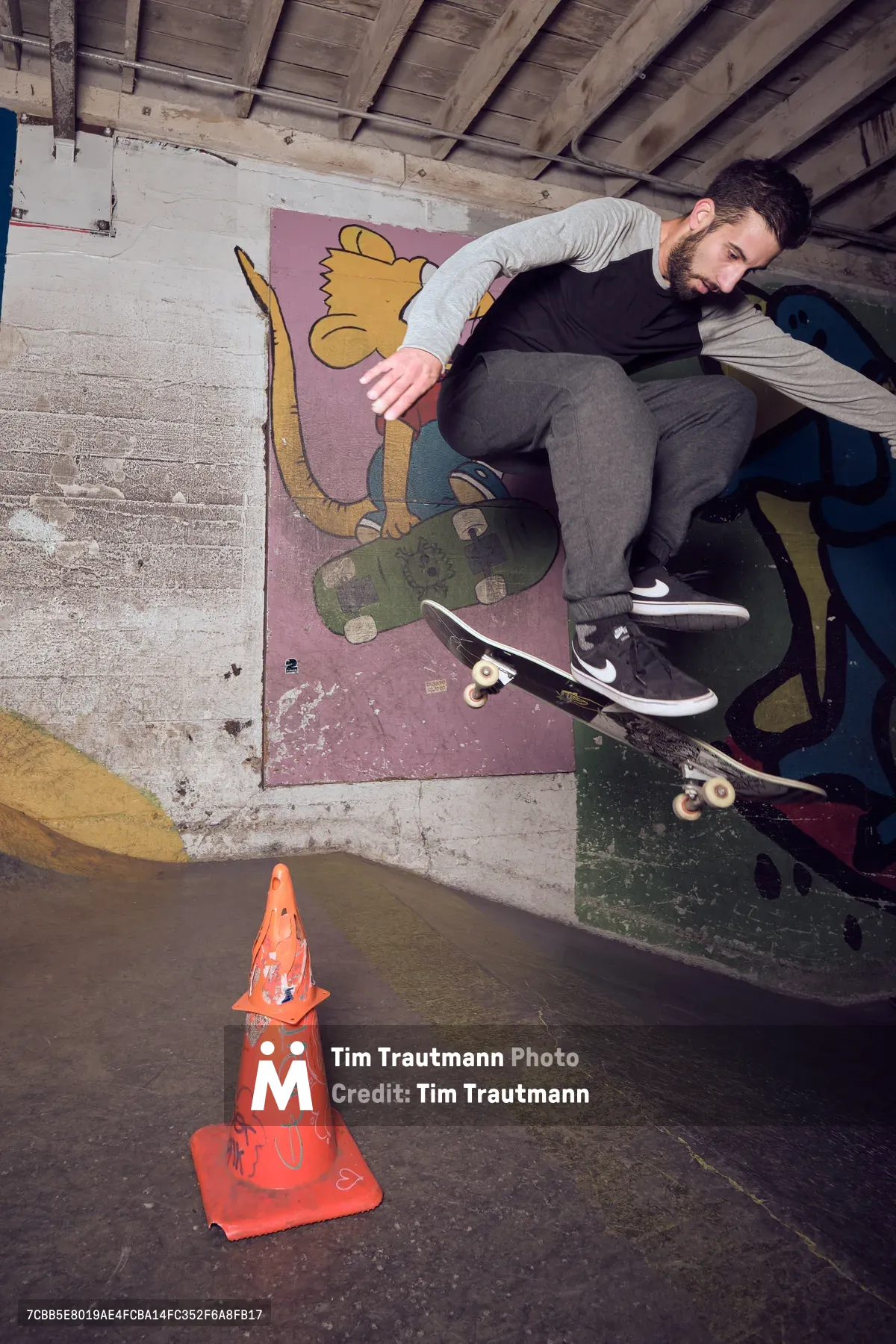 A bearded skateboarder in casual streetwear executes a wall ride against a vibrant mural in the raw concrete interior of Commonwealth Skateboarding in Portland's Buckman neighborhood. The industrial basement setting, marked by exposed wooden ceiling beams and weathered concrete walls, creates an authentic underground atmosphere. A spray-painted traffic cone sits prominently in the foreground, while colorful street art featuring a cartoon character adds urban energy to the gritty practice space.