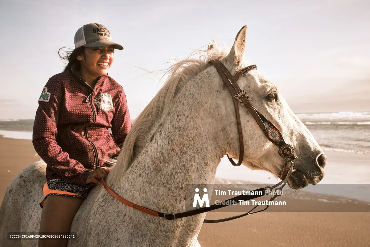 A radiant young woman in a burgundy polka-dot shirt and baseball cap shares an intimate moment with her dappled gray horse along the pristine coastline near Wheeler, Oregon. The golden hour light bathes both rider and mount in warm tones as ocean waves lap the sandy beach behind them. Her genuine smile and the horse's ornate bridle speak to a deep bond forged through countless rides along this rugged Pacific shoreline. The misty coastal atmosphere and expansive beach create a dreamlike backdrop for this portrait of equestrian partnership.
