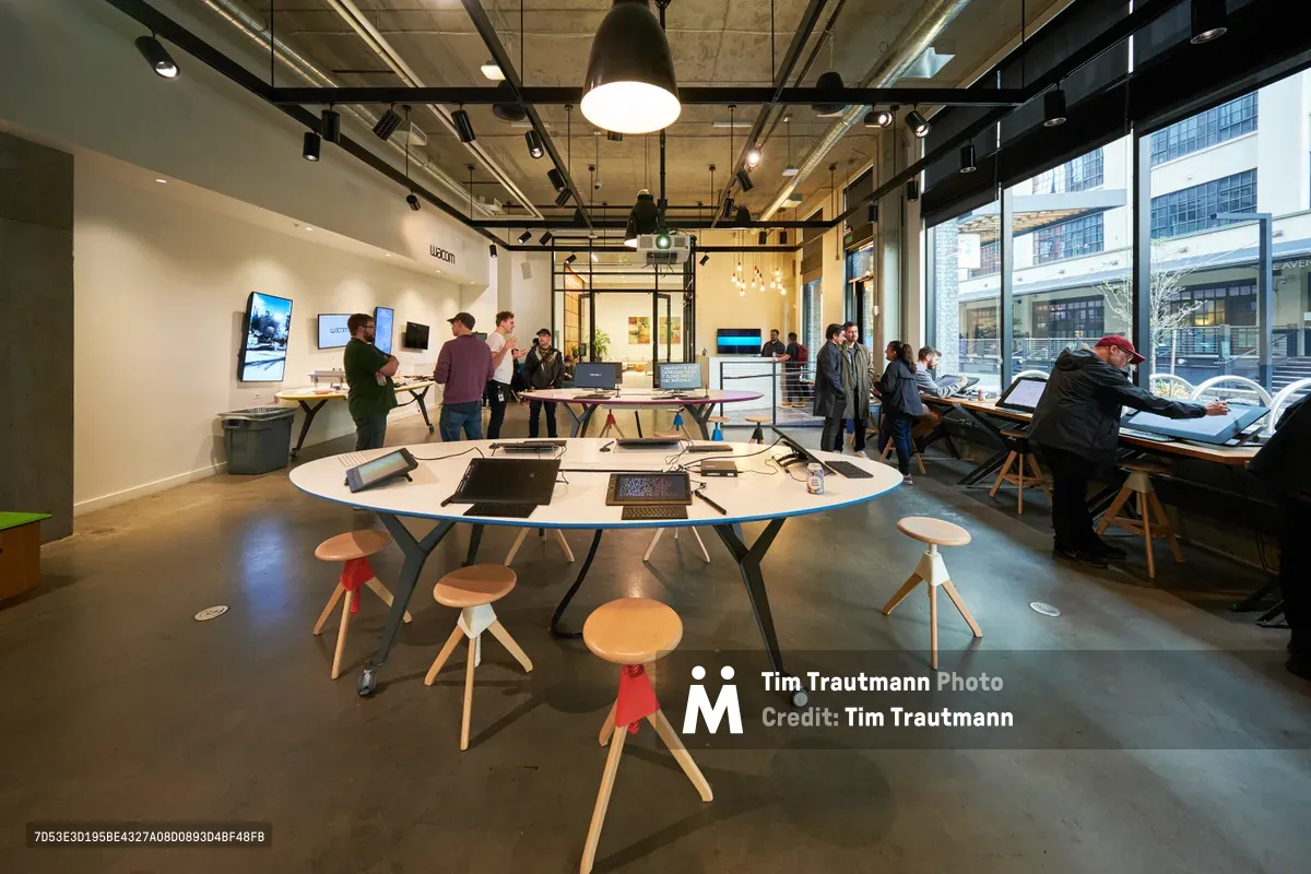 Inside the bright, industrial space of a modern technology center in Portland's Pearl District, creative professionals and enthusiasts gather around interactive workstations and digital displays. The exposed ceiling infrastructure and polished concrete floors create an urban workshop atmosphere, while large windows flood the space with natural light. Groups of people engage with digital devices at circular tables surrounded by colorful wooden stools, embodying the collaborative spirit of contemporary creative education.