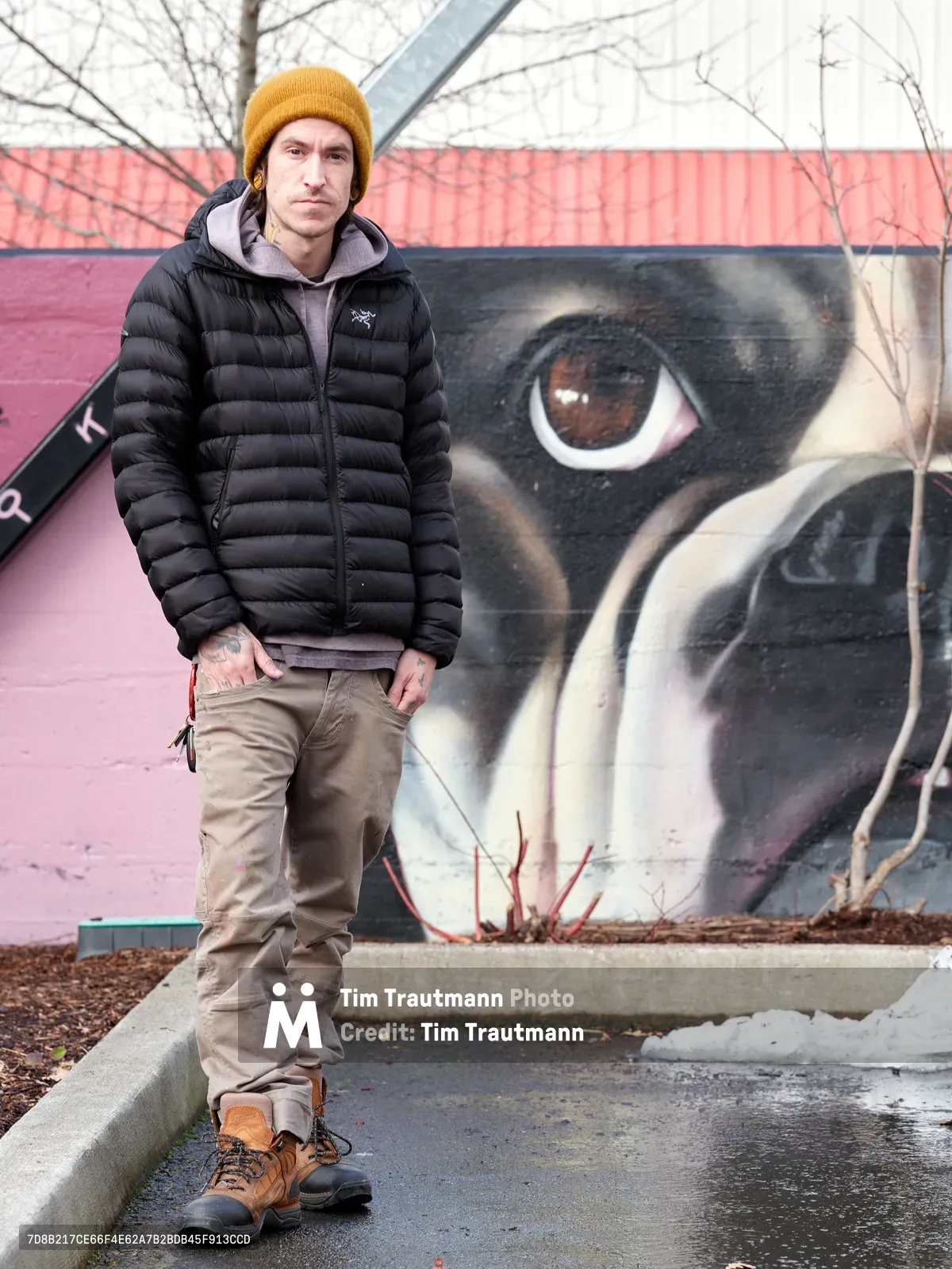 Muralist Wetiko stands confidently before his completed street art piece at the Clay Creative Building in Southeast Portland's Hosford-Abernethy neighborhood. Dressed in winter layers with a distinctive mustard beanie and black down jacket, the artist poses against his striking mural featuring an oversized eye rendered in dramatic blacks and earth tones. The late winter setting captures bare branches overhead and patches of melting snow, while the industrial creative space provides an authentic backdrop for this portrait of contemporary urban artistry.