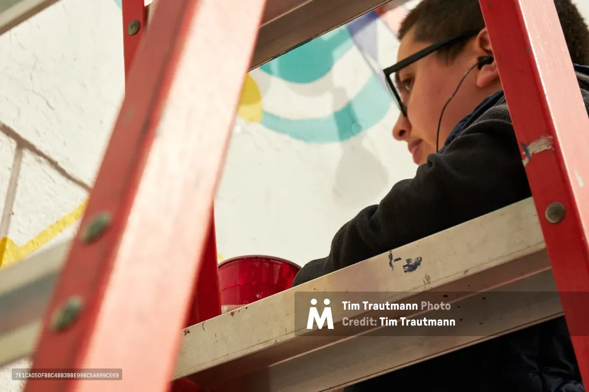Artist Anke Gladnick works intently on their mural commission "Let's Talk" within the creative spaces of Open Signal in Portland's Eliot neighborhood. Framed by coral-pink scaffolding and surrounded by paint supplies, the illustrator embodies focused artistic dedication as natural light illuminates their workspace. The scene captures the intersection of public art creation and community cultural programming in Northeast Portland's vibrant arts district.