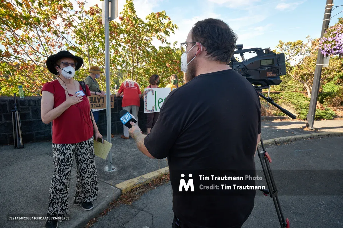 A masked climate activist in a red top and patterned pants speaks passionately to a KOIN Channel 6 news reporter during a rally outside Representative Kurt Schrader's Oregon City office. The cameraman positions his professional equipment to capture the interview as protesters with signs gather in the background under the dappled shade of autumn trees. The scene unfolds on McLoughlin Promenade, where civic engagement meets local journalism in the golden light of late afternoon.