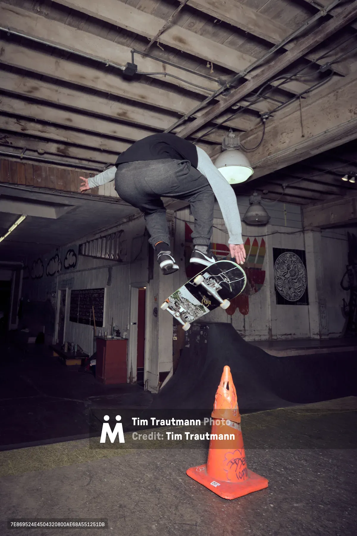 A skateboarder executes an airborne trick over a weathered orange traffic cone in the industrial interior of Commonwealth Skateboarding in Portland's Buckman neighborhood. The raw concrete floors and exposed ceiling beams create a gritty urban atmosphere, while graffiti and street art adorn the walls beneath harsh industrial lighting. The skater, captured mid-flight in dark clothing, demonstrates technical skill against the backdrop of this authentic underground skate venue.