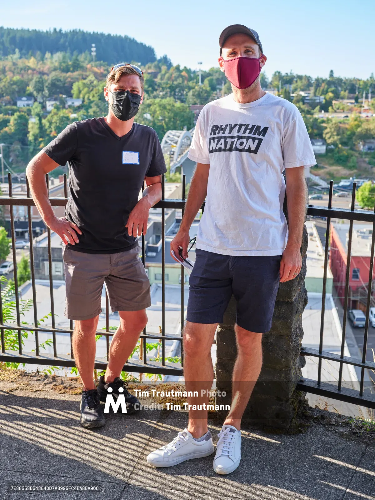 Michael Gaskill and Peter Marks stand side by side on McLoughlin Promenade overlooking Oregon City, both wearing protective masks during a climate change protest at Representative Kurt Schrader's office. The golden hour light casts dramatic shadows through metal railings as the forested hills and residential neighborhoods stretch into the distance. Gaskill wears a black v-neck with khaki shorts while Marks sports a white "Rhythm Nation" t-shirt and navy shorts, their relaxed poses contrasting with the serious political nature of their gathering.