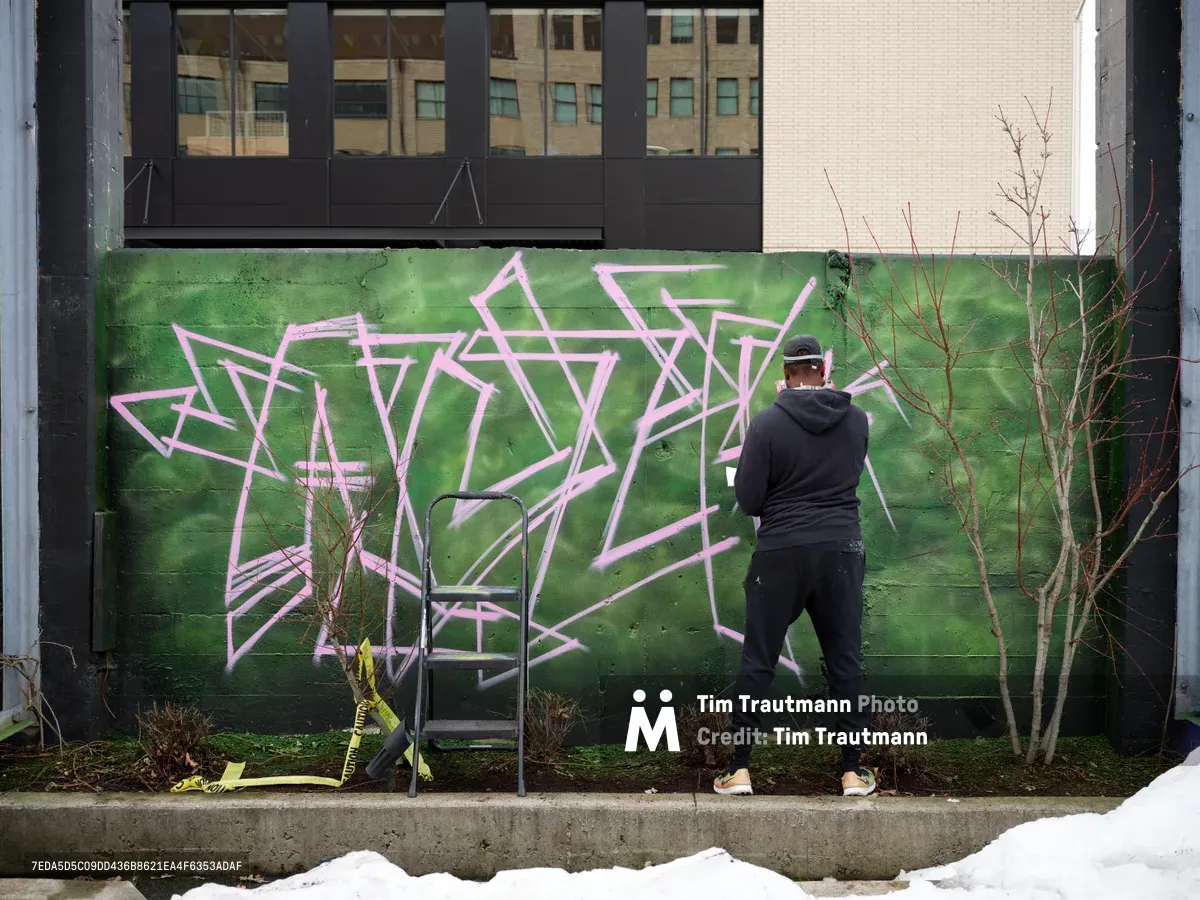 A hooded artist works methodically on an elaborate pink and white graffiti piece against a vibrant green wall in Southeast Portland's Hosford-Abernethy neighborhood. The scene unfolds in Clay Creative's parking lot, where winter-bare saplings frame the emerging artwork while urban architecture provides a backdrop of glass and brick. Yellow caution tape and construction materials hint at the ongoing transformation of this creative space.