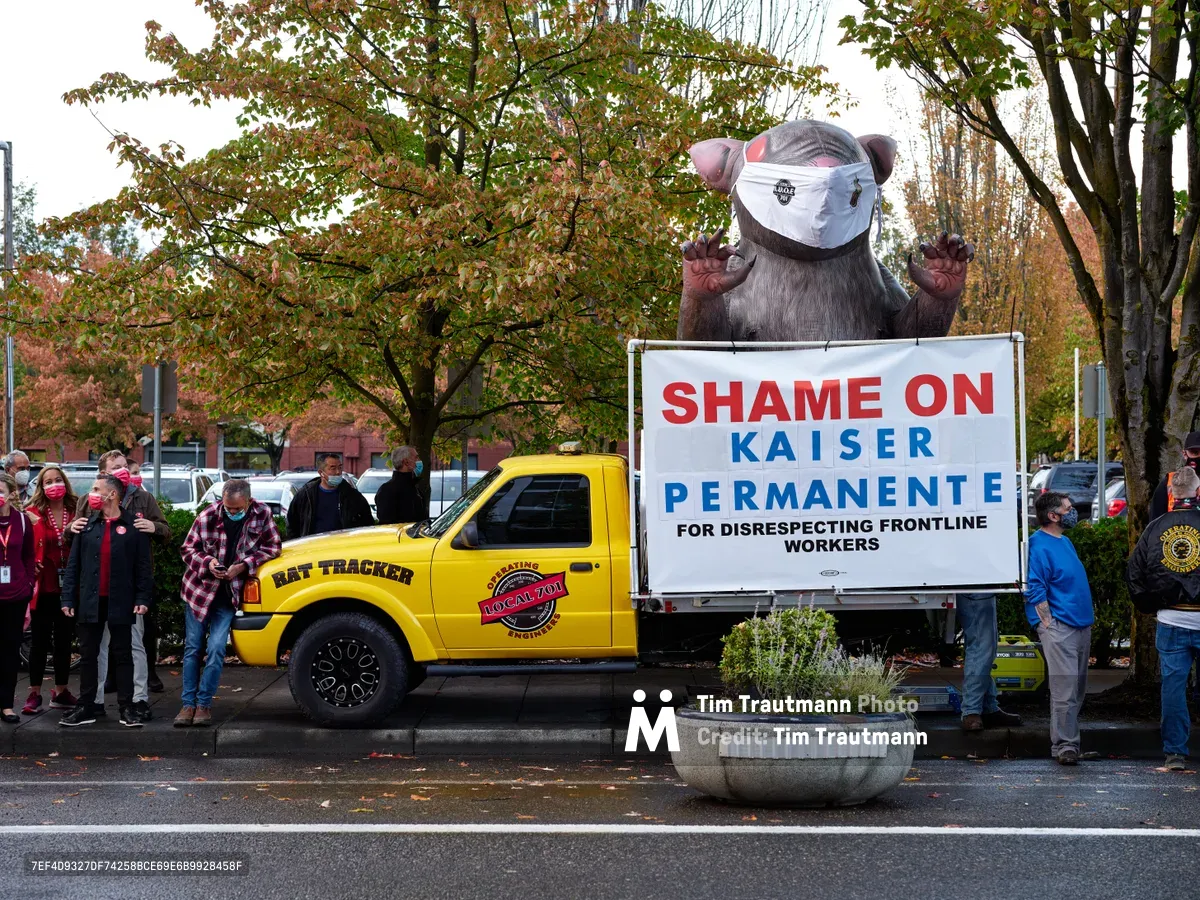 A towering inflatable rat wearing a surgical mask looms over a labor demonstration in Portland's Lloyd District, its claws raised menacingly beside a bright yellow truck bearing the message 'Shame on Kaiser Permanente.' Masked healthcare workers and union supporters cluster around the symbolic creature beneath autumn trees, their presence creating a powerful tableau of organized dissent. The overcast Oregon sky provides a muted backdrop to the bold red and blue protest signage, while fallen leaves scattered across the pavement hint at the season of change both literally and figuratively.