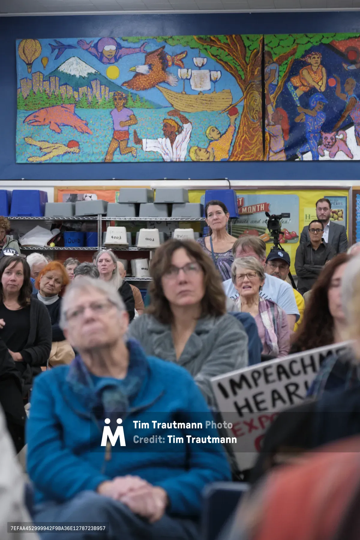 Beneath a vibrant community mural depicting Pacific Northwest landscapes and cultural themes, constituents gather in the cafeteria of Robert Gray Middle School in Portland's Hillsdale neighborhood for Senator Ron Wyden's town hall meeting. The assembled crowd of predominantly middle-aged and older adults reflects the engaged citizenry of Oregon, with one attendee prominently holding an impeachment hearing sign. The institutional fluorescent lighting and casual school setting create an intimate, grassroots atmosphere where democratic participation unfolds against the backdrop of colorful artwork celebrating regional identity.