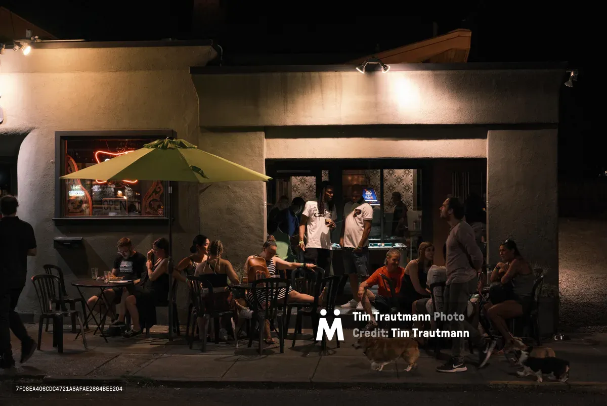 Warm tungsten light spills from Binks cafe onto Northeast Alberta Street during Portland's Last Thursday street fair, illuminating clusters of diners savoring the late evening atmosphere. Groups of friends occupy small tables beneath a sage-colored umbrella while others stand in animated conversation, their silhouettes softly defined against the glowing storefront windows. The intimate sidewalk scene captures the organic social rhythms of this monthly community celebration in the Concordia neighborhood.