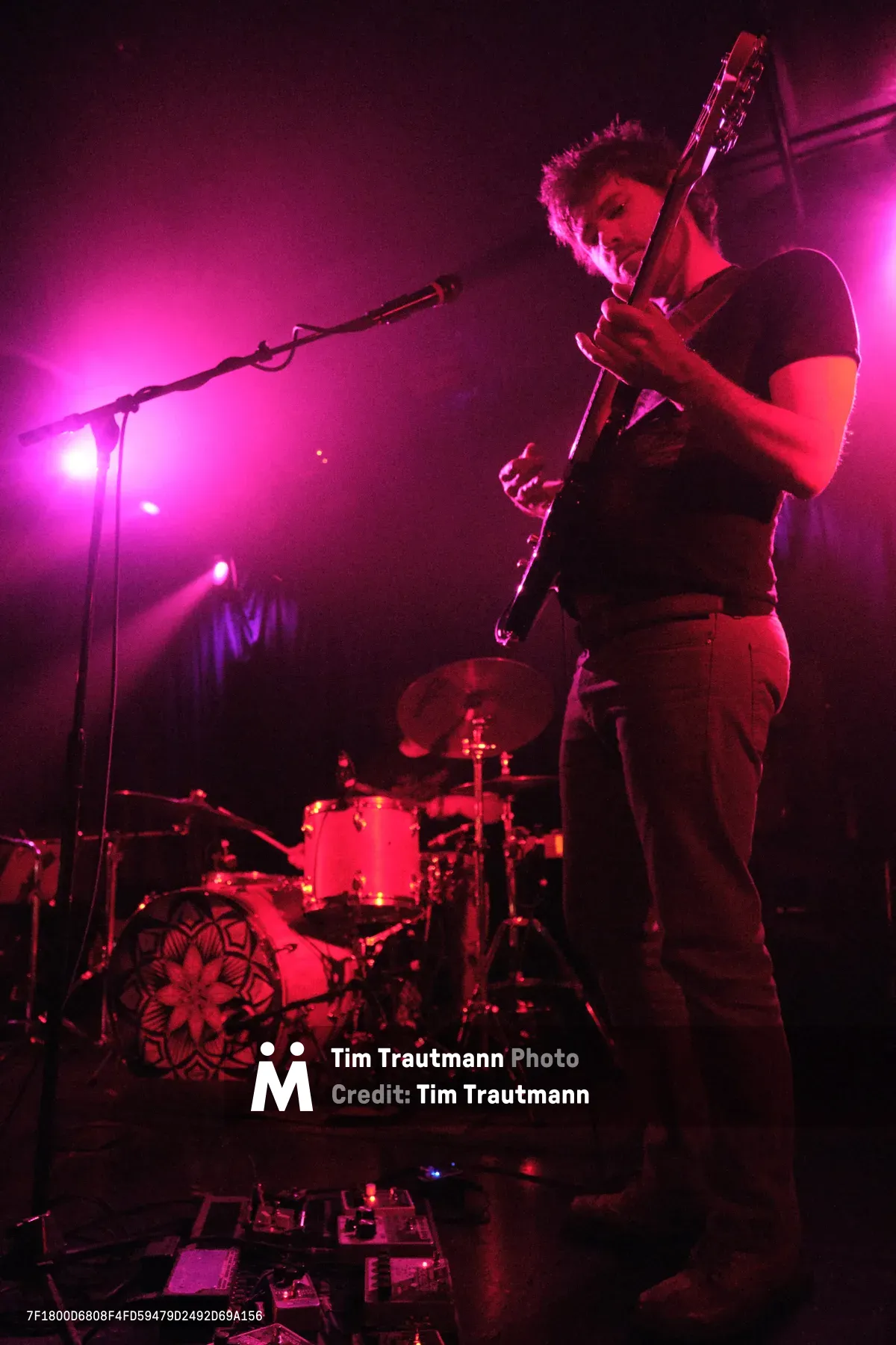 A guitarist from Sun Blood Stories commands the stage at Dante's in downtown Portland, bathed in deep magenta and red stage lighting that creates an otherworldly atmosphere. The musician, dressed in casual dark clothing, grips an electric guitar while positioned near the microphone stand, with a drum kit visible in the atmospheric haze behind him. The intimate venue setting captures the raw energy of live music performance, where dramatic lighting transforms the mundane into the cinematic.