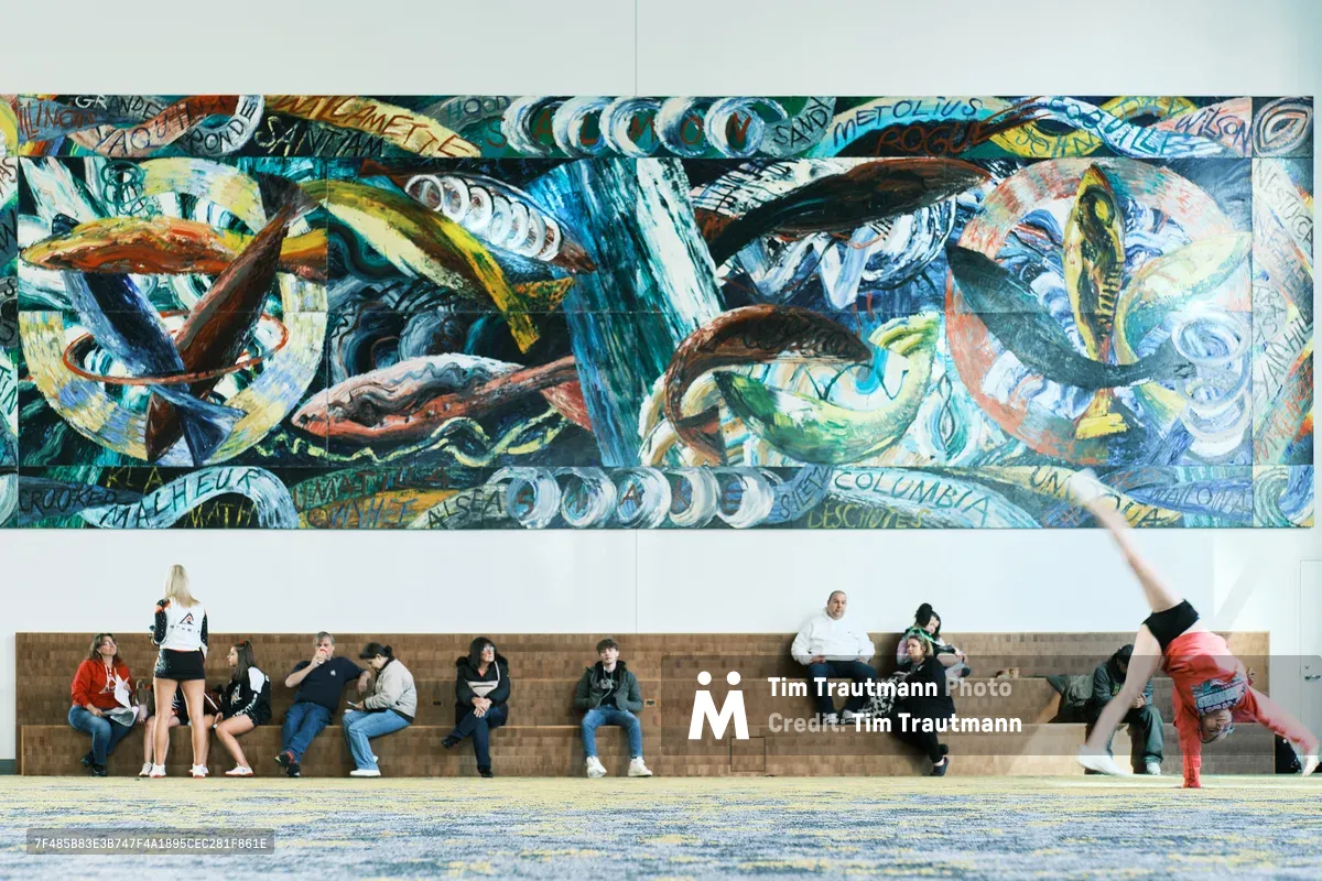 Visitors seek momentary refuge on stone benches beneath an explosive panorama of maritime-themed murals at Portland's Oregon Convention Center. The vibrant wall art, dominated by swirling blues and oceanic motifs interwoven with text fragments, creates a dramatic backdrop for the diverse group of attendees—from a breakdancer frozen mid-move to couples and individuals in casual repose. The juxtaposition of kinetic street art energy above and human stillness below captures the convention center's role as both cultural crossroads and temporary sanctuary.