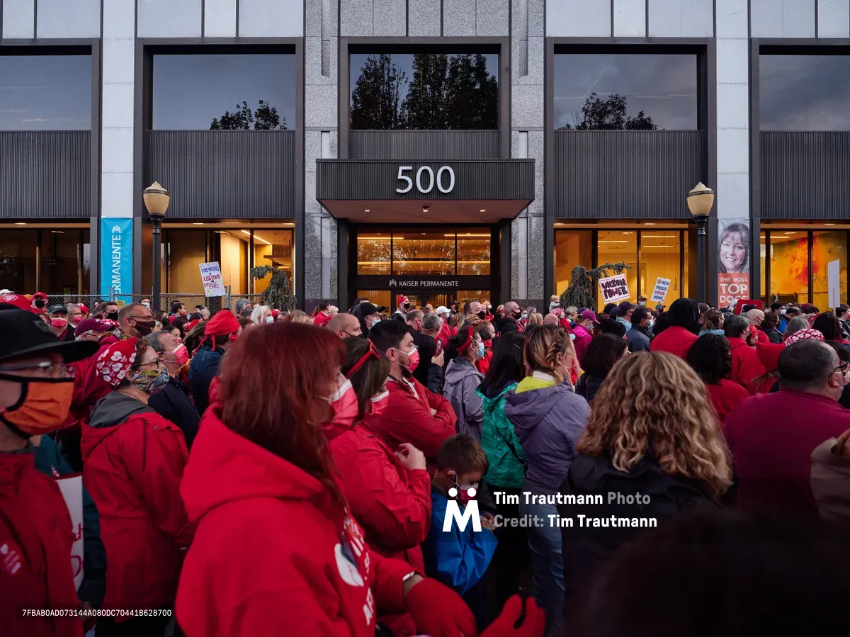A dense crowd of healthcare workers, predominantly wearing red Kaiser Permanente scrubs and winter jackets, gathers in solidarity outside the Kaiser Permanente Tower at 500 Northeast Multnomah Street in Portland's Lloyd District. The sea of crimson creates a powerful visual statement against the modern glass and steel facade of the medical facility, while protest signs punctuate the crowd and evening light casts a determined atmosphere over the assembled nurses. Face masks worn by many participants underscore the contemporary healthcare context of their labor action.