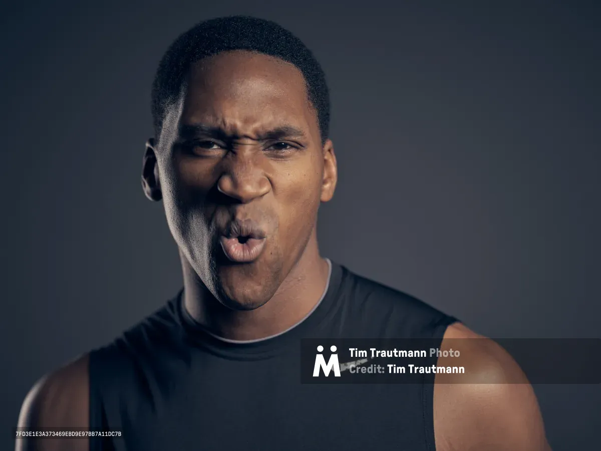 A powerful studio portrait captures a young Black athlete mid-expression, his face contorted with intense focus and determination. Dramatic lighting sculpts his features against a moody gray backdrop, while his black athletic shirt emphasizes his athletic build. The image demonstrates the exceptional detail and tonal range capabilities of medium format photography, shot during a camera system evaluation in Portland's Northwest District.