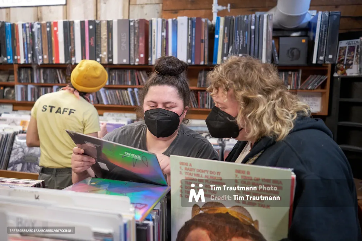 Two masked women lean intimately over a collection of vinyl records at Music Millennium on East Burnside Street during Record Store Day 2022 in Portland, Oregon. The brunette staff member with a top knot and the curly-haired blonde customer share a moment of musical discovery, examining album covers amid towering shelves of CDs and records. Warm indoor lighting illuminates this authentic record store scene, while a staff member in a yellow beanie browses in the soft-focused background.