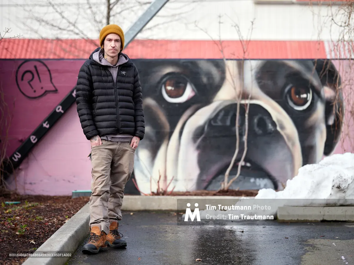 Painter and muralist Wetiko stands confidently before his striking dog portrait mural at Clay Creative Building in Southeast Portland's Hosford-Abernethy neighborhood. The artist wears a golden mustard beanie and black puffer jacket against the late winter atmosphere, with patches of snow visible on the concrete. Behind him, his hyperrealistic mural depicts a close-up of a dog's face with expressive brown eyes and detailed fur work, painted in warm tones against the building's coral-pink exterior wall.