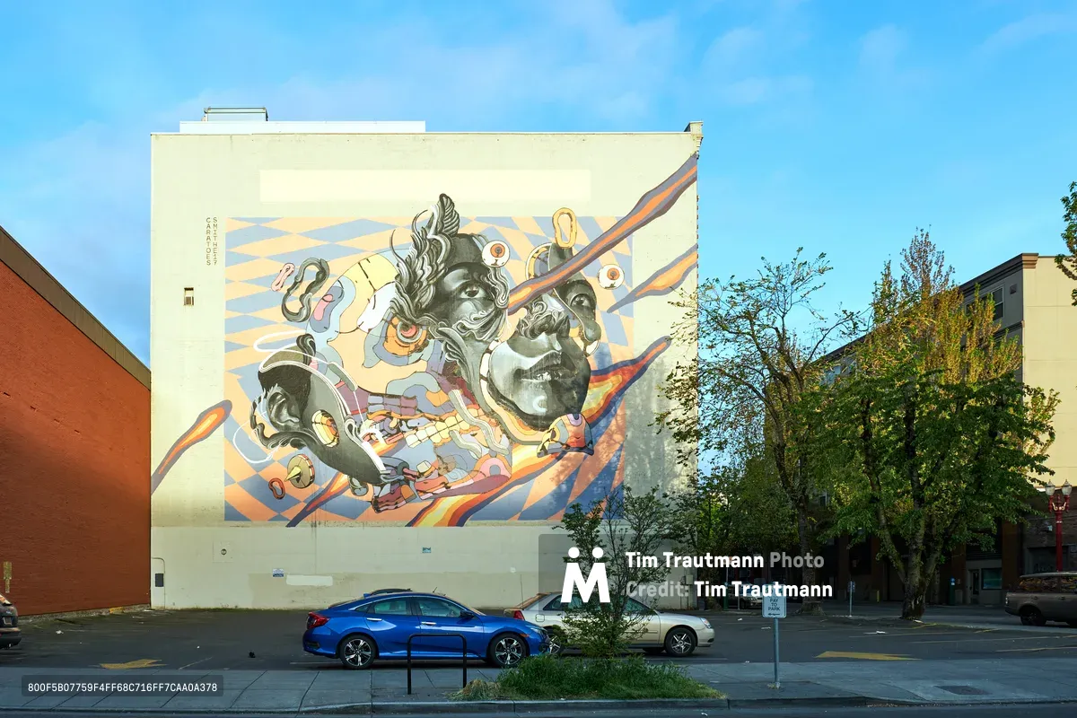 A monumental street mural dominates a white building wall in Portland's historic Chinatown district, featuring stylized portraits rendered in flowing ribbons of coral, blue, and gold against geometric patterns. The artwork depicts figures with distinctive Asian features surrounded by swirling decorative elements, creating a dreamlike narrative that bridges cultural identity and contemporary urban art. Blue and white sedans rest quietly along the tree-lined street below, while spring foliage frames the scene under an expansive blue sky dotted with wispy clouds.