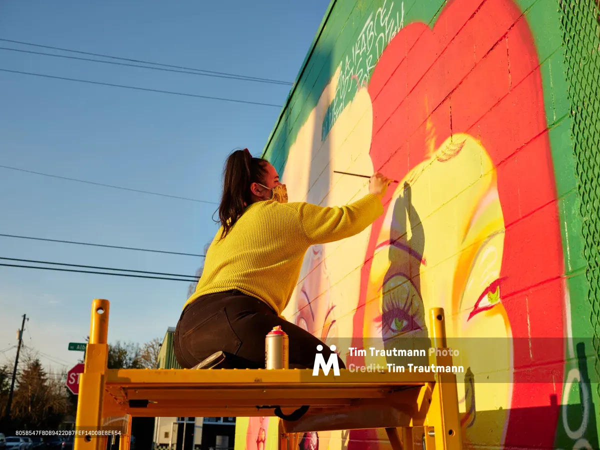Multidisciplinary artist Villatose works from a wooden scaffolding platform, adding delicate brushstrokes to her large-scale mural 'Jardín Cubana' depicting three female figures on Northeast Alberta Street in Portland's celebrated arts district. Golden hour light bathes the scene, illuminating her mustard yellow sweater against the mural's bold palette of coral, emerald, and sunshine yellow. Power lines stretch across the azure sky above, while spray paint cans rest on the platform beside her as she brings life to the colorful portrait emerging on the building's facade.