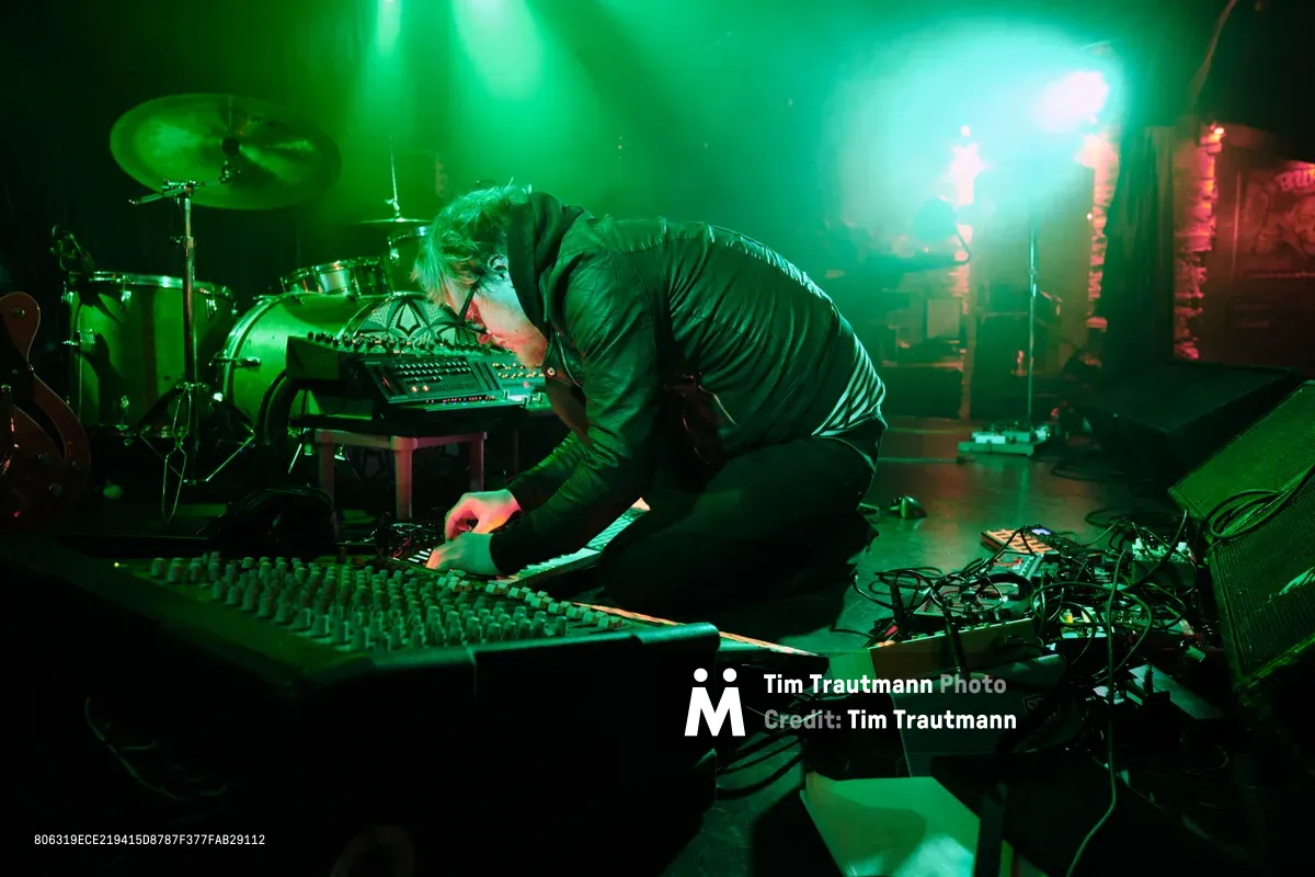 Electronic musician Eli Savage, performing as ellipsism, hunches intensely over a mixing console amid a sea of synthesizers and equipment at Portland's legendary Dante's venue. Ethereal green stage lighting bathes the intimate performance space, creating an otherworldly atmosphere as the artist manipulates sound through an array of electronic instruments. Behind him, a drum kit and additional gear fade into the moody shadows, while tangled cables snake across the stage floor, evidence of the complex sonic architecture being constructed in real time.