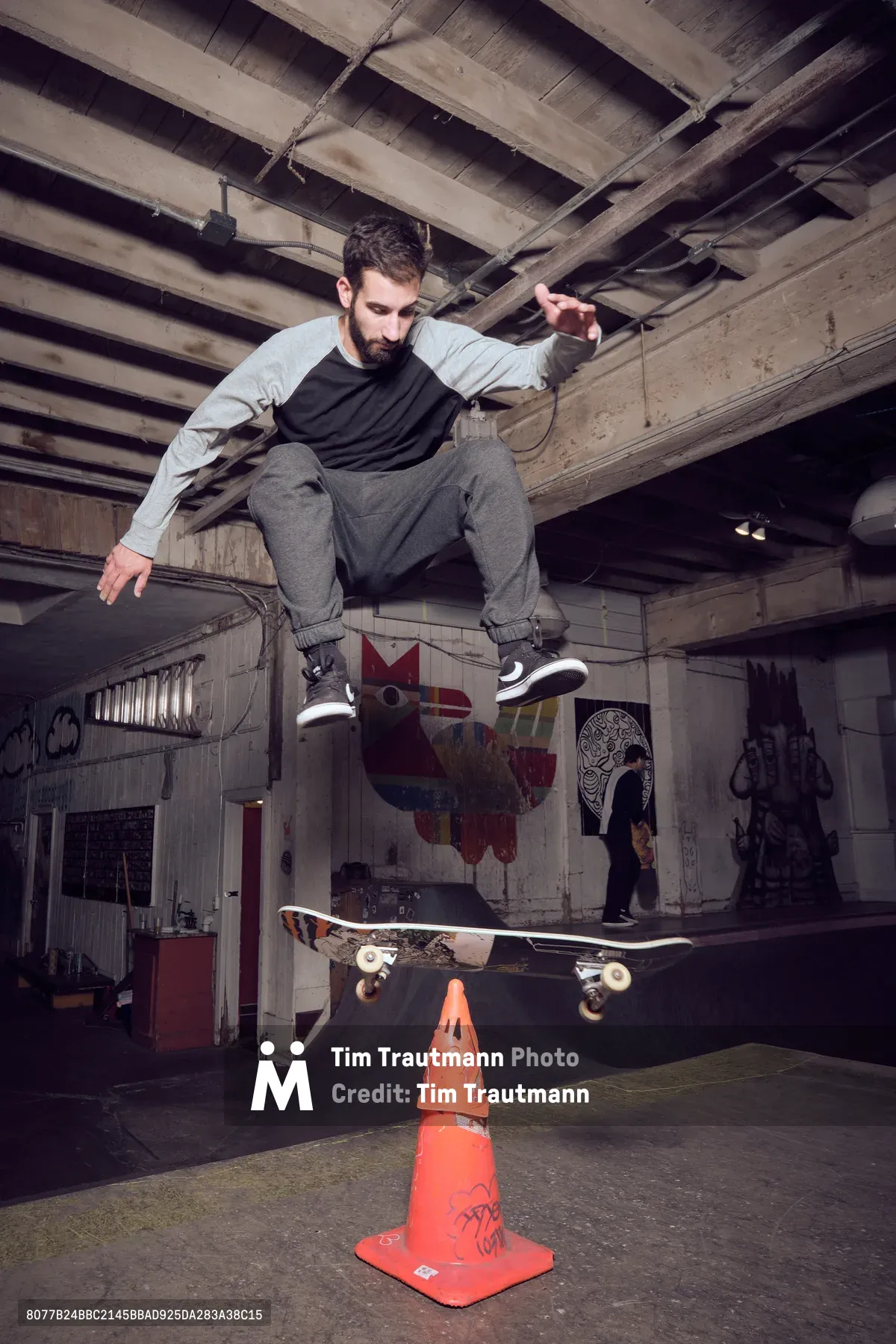 A bearded skateboarder in grey sweats and black tee defies gravity in the raw concrete underbelly of Commonwealth Skateboarding in Portland's Buckman neighborhood. The industrial space's exposed wooden ceiling beams frame this moment of athletic suspension as he soars above a weathered orange traffic cone, his skateboard floating beneath him. Graffiti-tagged walls and shadowy recesses create an authentic urban backdrop for this editorial skateboarding portrait.