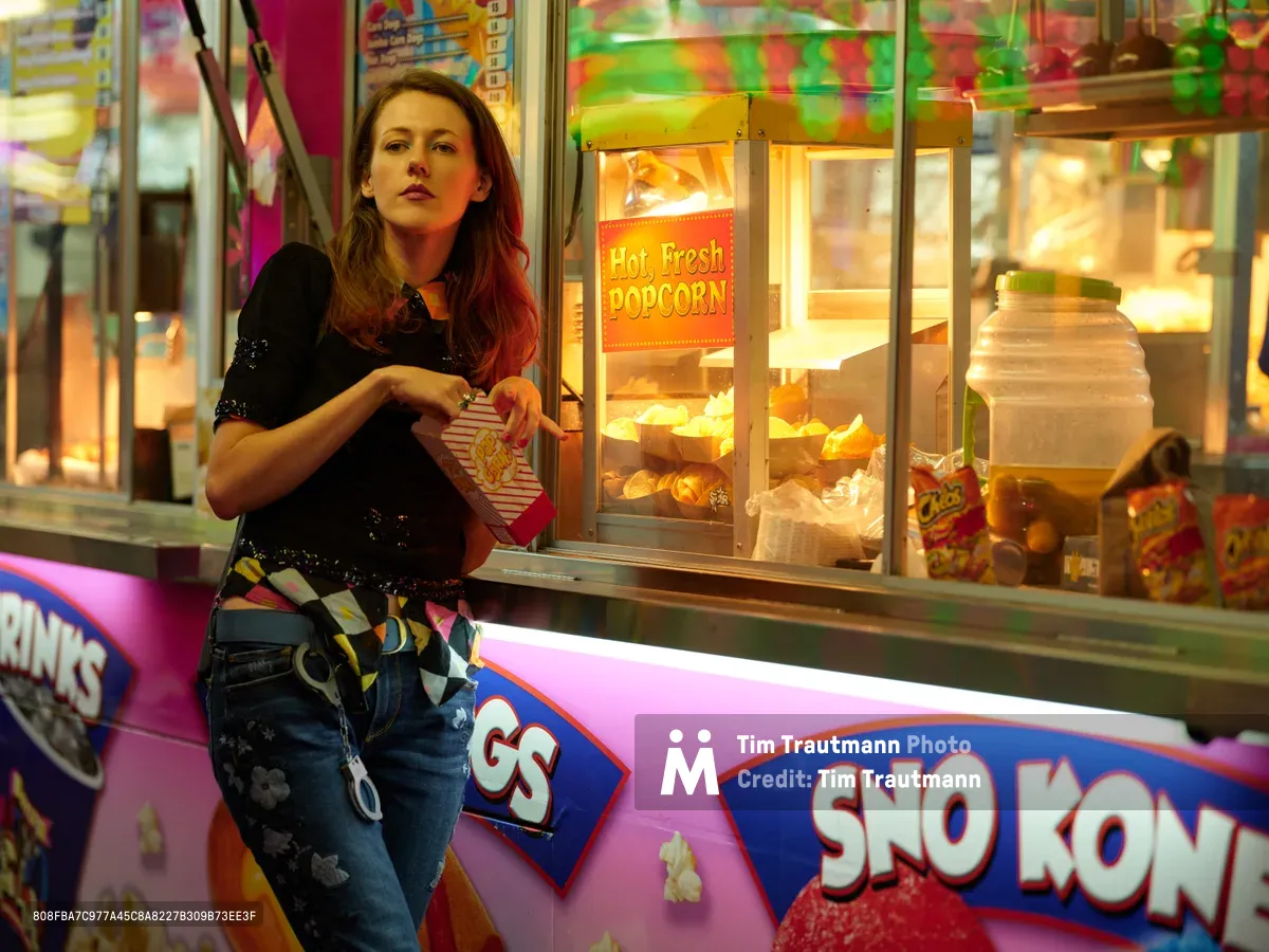 A young woman in distressed denim and a sequined black top leans against a vibrant concession stand at the Clark County Fair in Vancouver, Washington. The golden glow from the popcorn machine illuminates her contemplative expression as neon carnival lights create a dreamy bokeh in the background. The scene captures the nostalgic magic of fairground evenings, where the aroma of kettle corn mingles with the electric energy of carnival attractions.