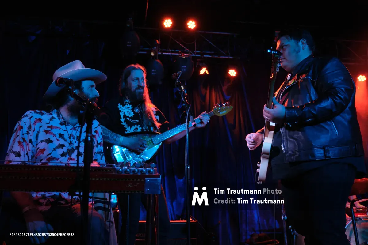 Barry Walker in a cowboy hat and floral shirt commands the keyboards while Tobias Berblinger and Victor Franco flank him with guitar and bass in the atmospheric lighting of Dante's club. Crimson stage lights pierce through the intimate venue's darkness, casting the Portland trio in warm amber and electric blue hues as they deliver their performance during RX Fest 2019. The cramped stage setup and close quarters create an electric intimacy between the musicians and their audience.