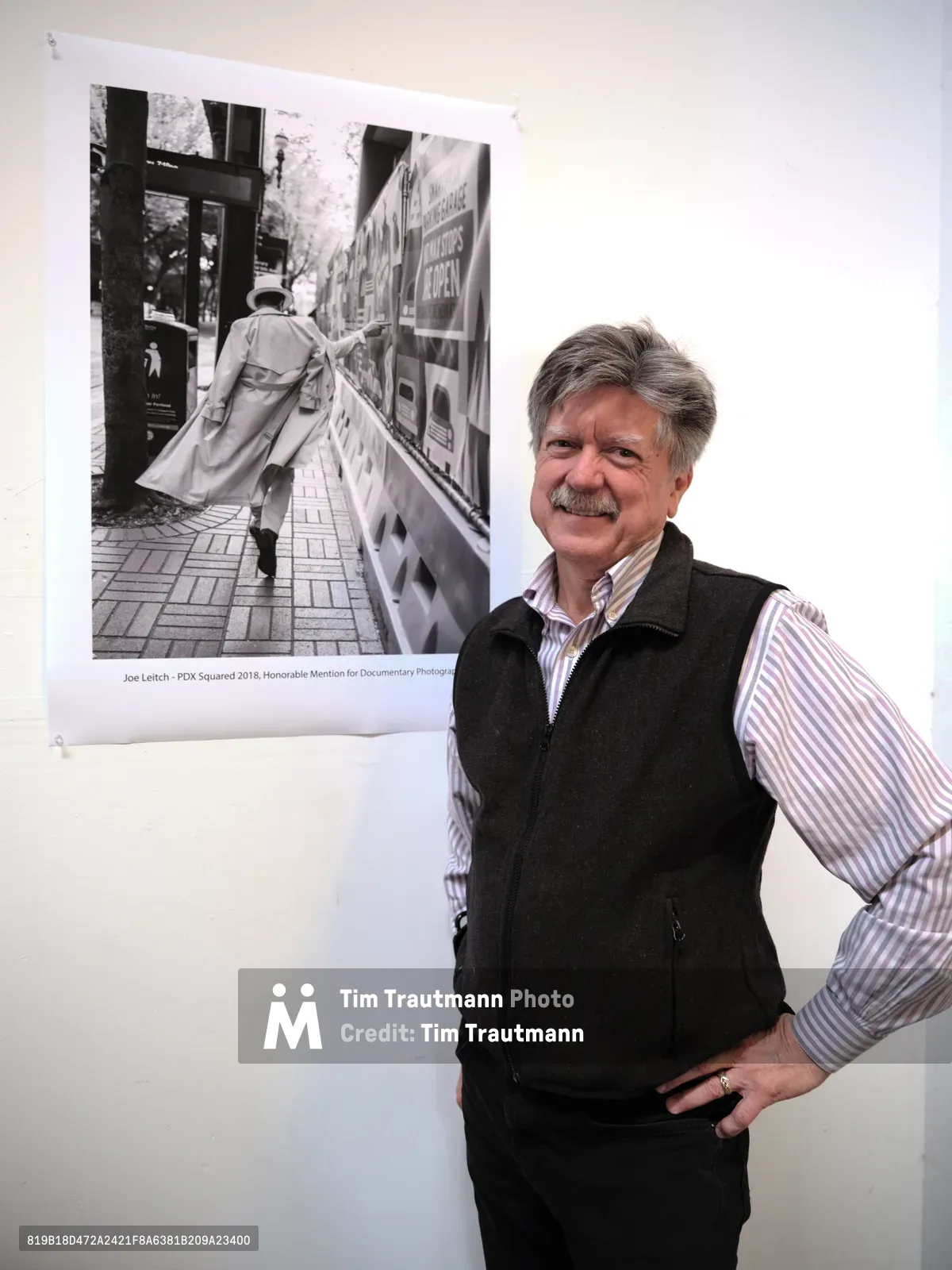 Joel Leitch, last year's documentary category winner, stands proudly beside his award-winning black and white street photograph at ASMP Oregon's PDX Squared 2019 exhibition in Portland. The mature photographer, wearing a black vest over a striped shirt, poses with confidence next to his compelling urban scene depicting a trench-coated figure walking past newspaper stands. The gallery's pristine white walls provide stark contrast to the gritty documentary aesthetic of his displayed work, creating a moment where artistic achievement meets humble presentation.