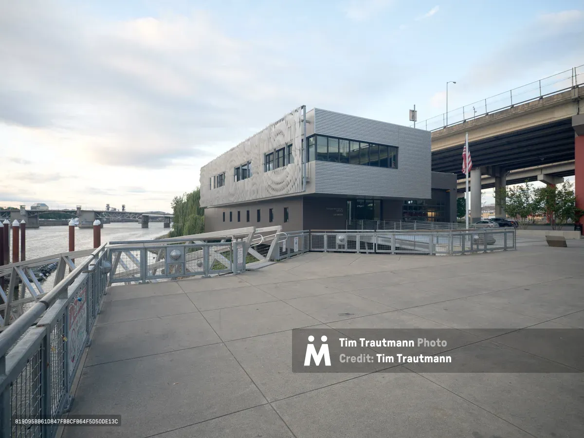 Portland Fire & Rescue Station 21 stands as a striking example of contemporary civic architecture along the Willamette River waterfront. The three-story building features a distinctive white sculptural facade contrasting with corrugated metal siding, positioned beneath the Southeast Madison Street bridge structure. Concrete walkways with metal railings frame the foreground under an overcast evening sky, while the distant river and bridge create layers of urban infrastructure.