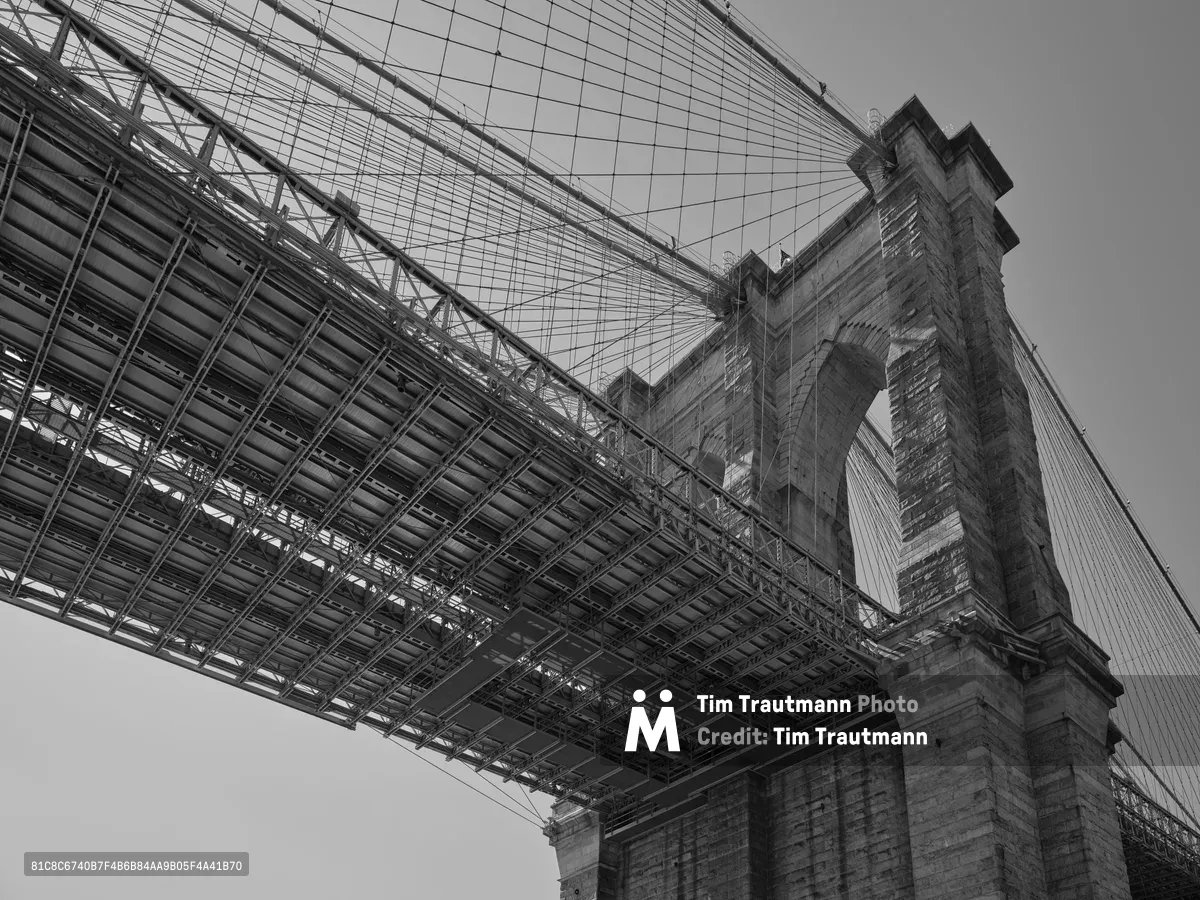 A dramatic worm's-eye view captures the Gothic Revival stonework of the Brooklyn Bridge's tower through the intricate steel lattice of the bridge deck structure above. Shot in monochrome from Emily Warren Roebling Plaza in DUMBO, the composition emphasizes the engineering marvel's dual nature - the romantic masonry arches juxtaposed against the industrial precision of steel cables and support beams. The low angle transforms the familiar landmark into an abstract study of intersecting lines and geometric patterns, with the tower's crenellated crown piercing the overcast Brooklyn sky.