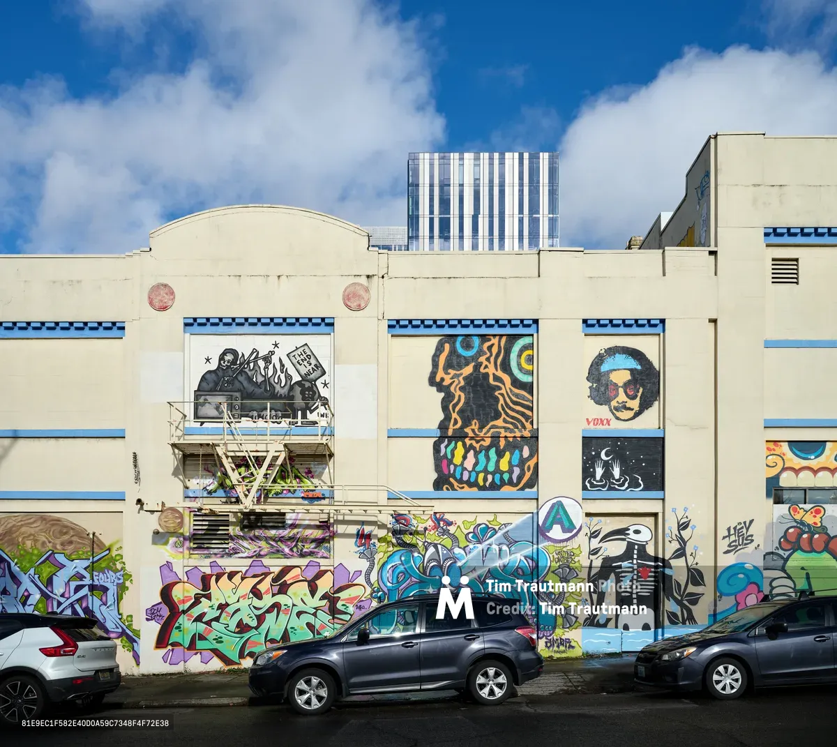 A vibrant explosion of commissioned street murals adorns the cream-colored façade of a former Salvation Army warehouse in Portland's Central Eastside district. The artwork features bold characters including a skeletal figure, psychedelic faces, and colorful graffiti-style lettering that cascades across multiple building sections. Two parked SUVs sit beneath the towering wall art against a backdrop of blue sky dotted with white cumulus clouds, while modern high-rise buildings pierce the horizon beyond.