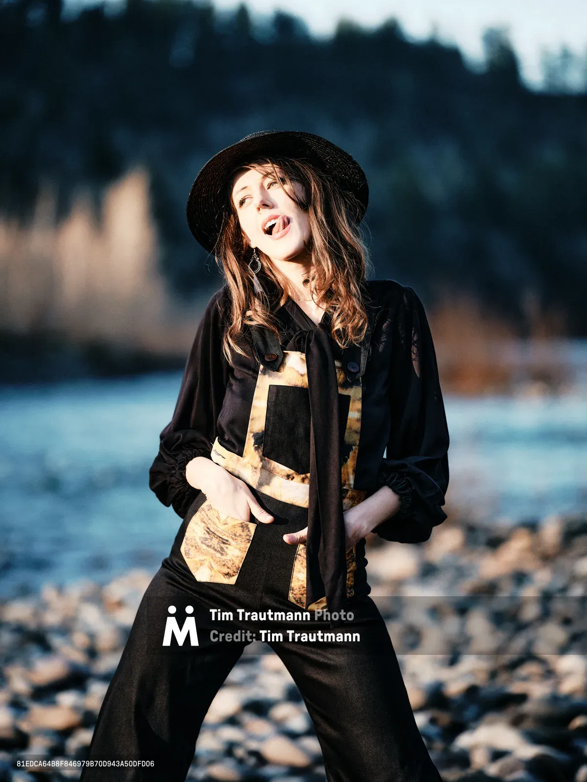 A young woman radiates pure joy as golden hour light illuminates her face along the rocky riverbed of Oregon's Floodplain Trail in Multnomah County. She wears a black wide-brimmed hat and layered earth-toned clothing, her mouth open in mid-laugh as she poses playfully against the backdrop of the Columbia River and distant forested hills. The warm, directional sunlight creates dramatic contrast between her sunlit figure and the soft blue tones of the water and sky behind her.