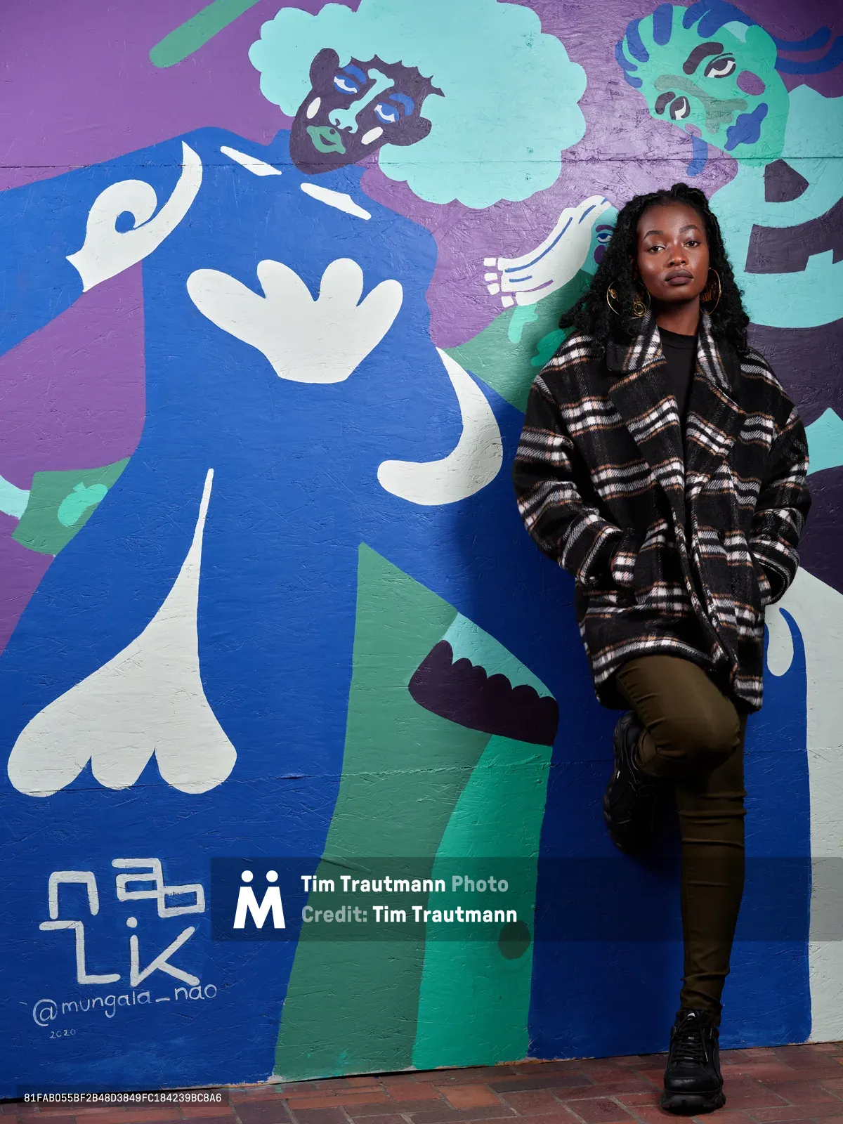 Naomi Likayi stands confidently against her vibrant street art mural painted on the boarded-up Portland World Trade Center, commissioned by the Portland Street Art Alliance. The contemporary artwork features bold blues, purples, and mint greens with abstract faces and flowing forms, creating a striking backdrop for the artist's portrait. Likayi wears a black and white plaid flannel over olive pants, her locs framing her face as she poses with quiet authority beside her public art creation.