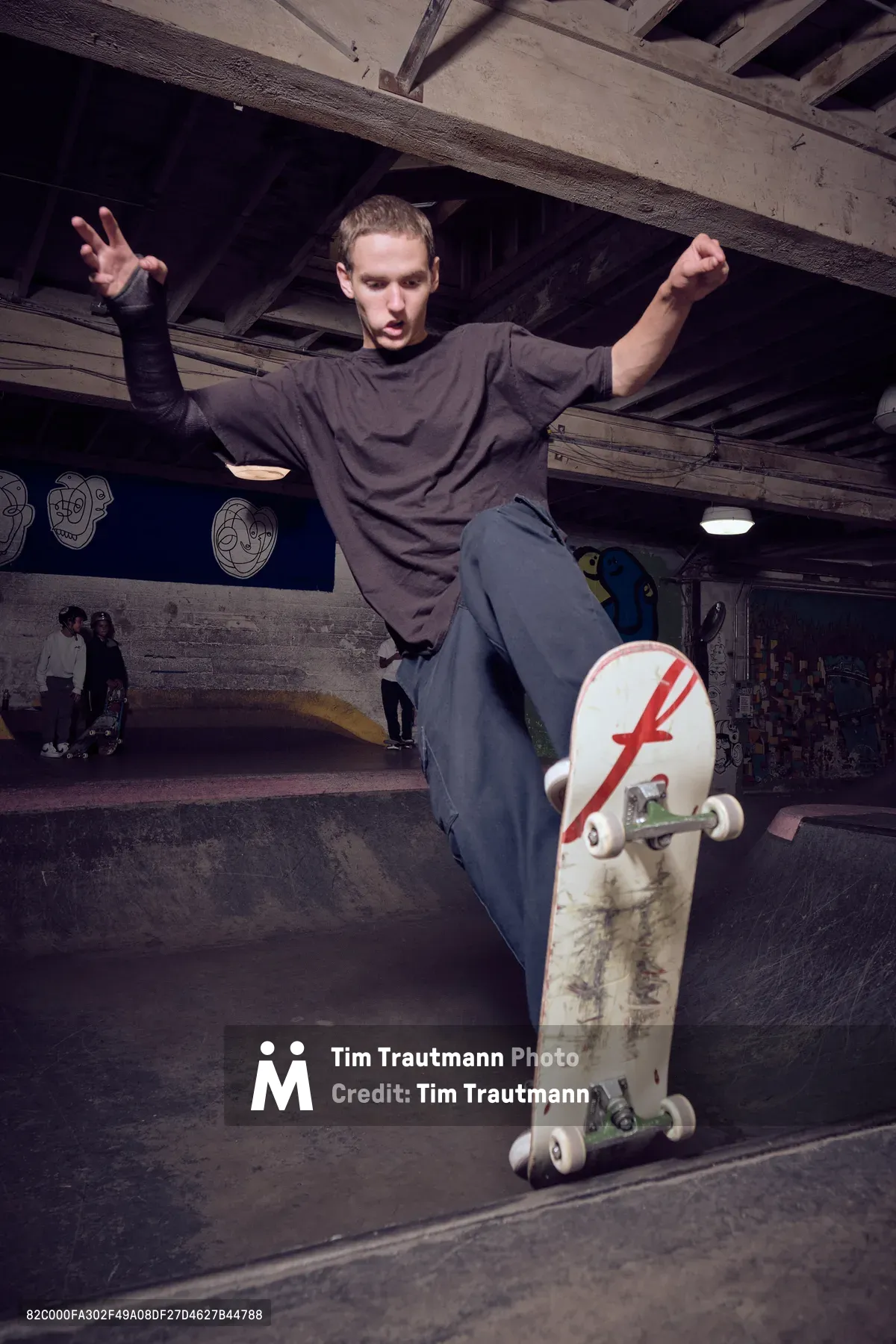 A young skateboarder carves through the curved concrete bowl at Commonwealth Skateboarding in Portland's Buckman neighborhood, his arms outstretched for balance as his weathered deck cuts through the transition. The industrial ceiling beams and graffitied walls create a raw urban backdrop, while fellow skaters occupy the background shadows. Warm artificial lighting illuminates the action, capturing the fluid motion and concentrated expression of street skateboarding culture.