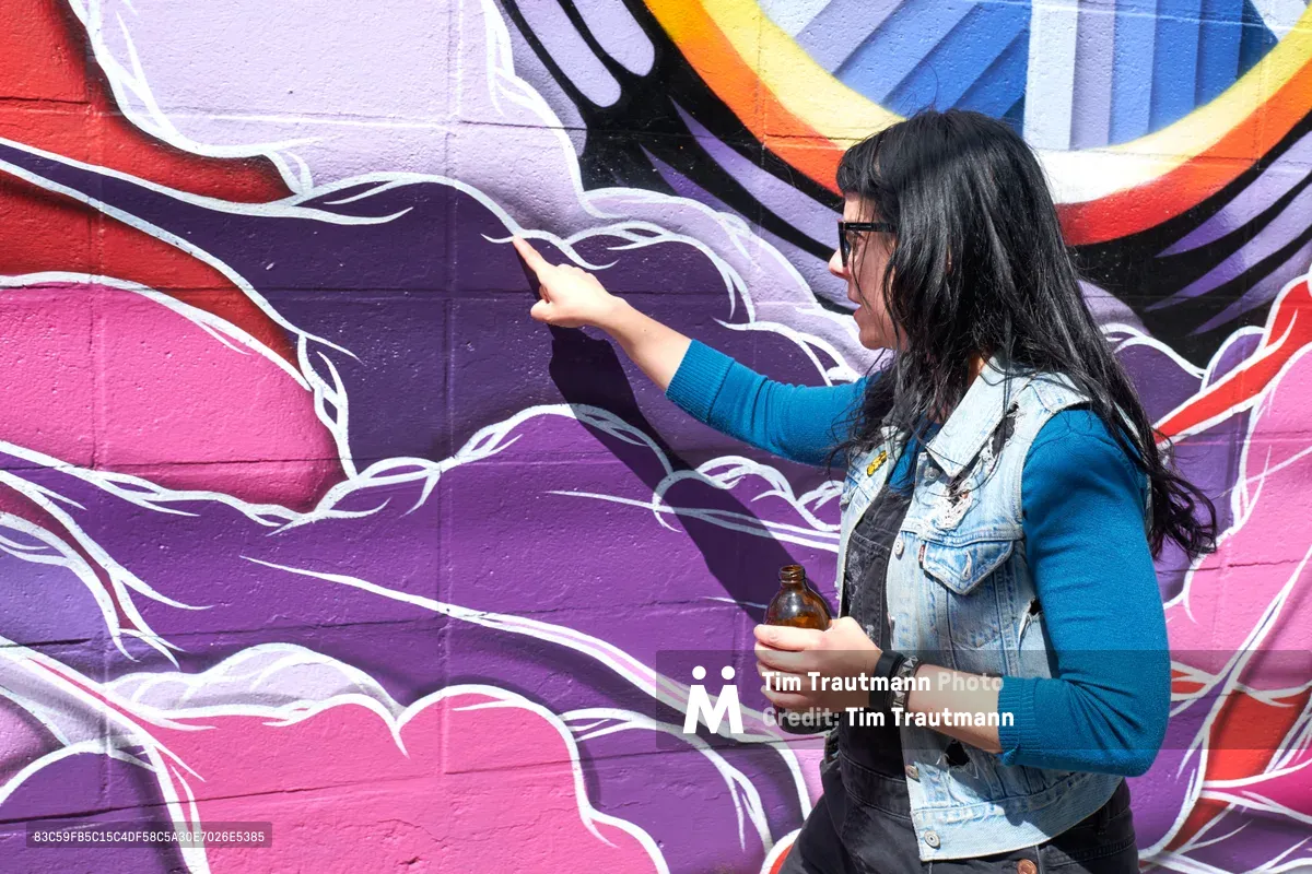 Tiffany Conklin of the Portland Street Art Alliance gestures toward a vibrant purple and pink mural while leading an interpretive tour in Portland's Central Eastside neighborhood. Dressed in a blue sweater and denim vest, she holds a bottle while explaining the artwork's significance against the backdrop of flowing, organic forms painted in deep purples, magentas, and warm orange accents. The scene captures the intimate, educational nature of community-led cultural tours.